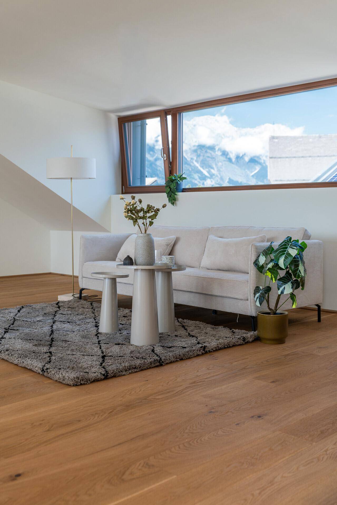 Gemütliches Wohnzimmer mit hellem Sofa, Holzboden und einem großen Fenster mit Bergblick.
