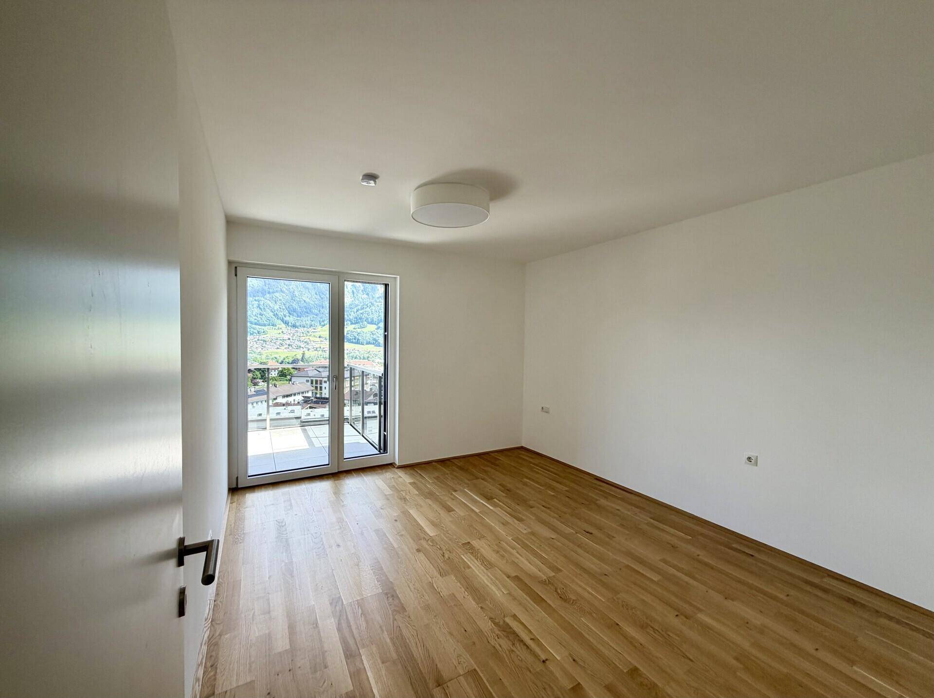 Schlafzimmer mit hellem Holzparkett, großen Fenstern und direktem Zugang zu einem Balkon mit Bergblick.