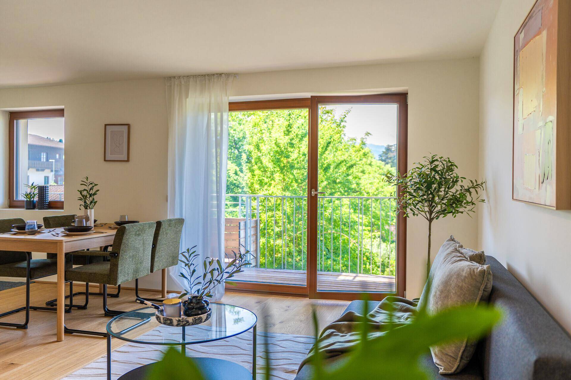 Heller Wohn- und Essbereich mit Holzboden, großen Fenstern und Zugang zum Balkon mit grüner Aussicht.