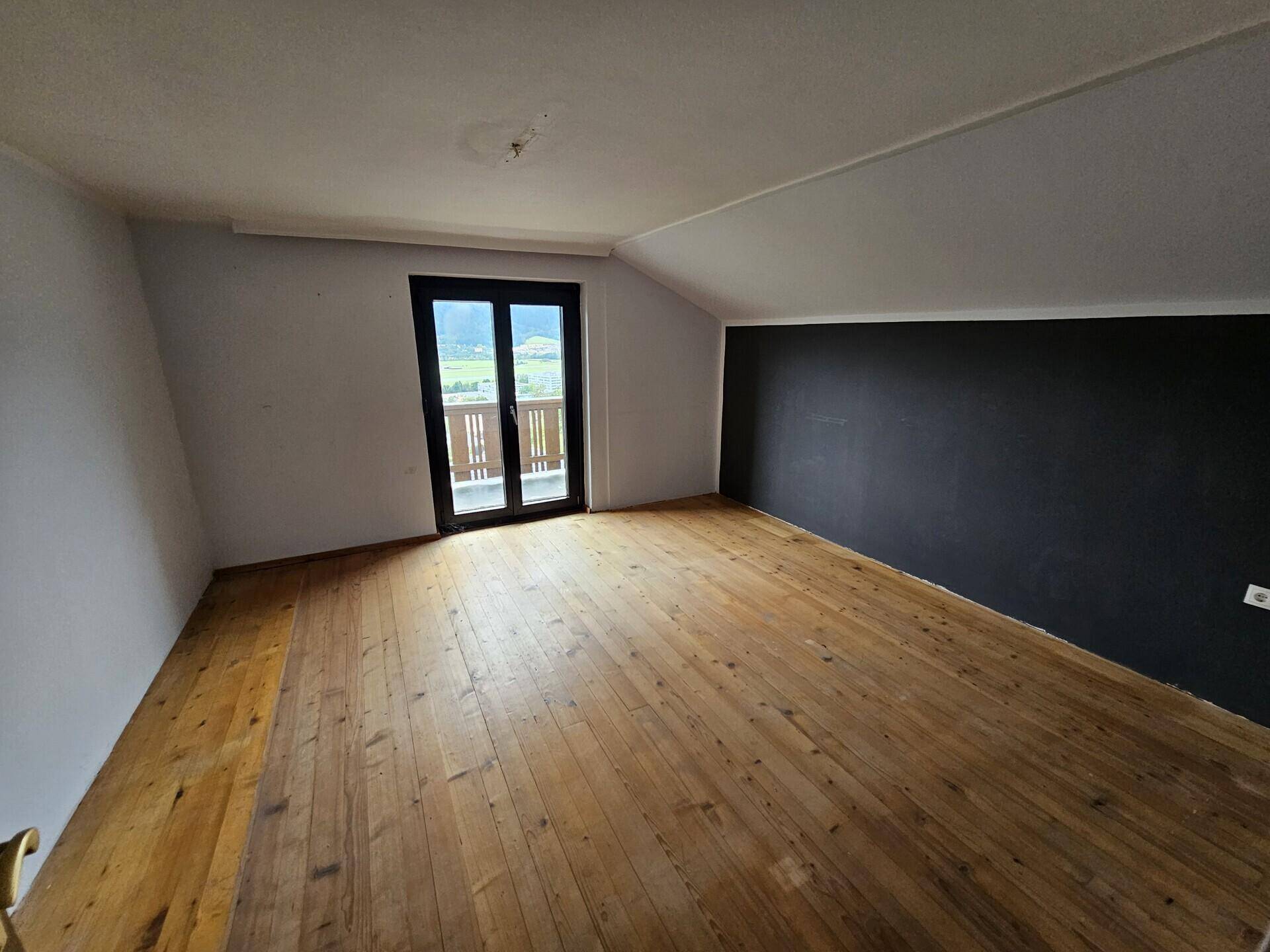 Geräumiges Dachzimmer mit Holzboden, dunkler Akzentwand und Balkonzugang.