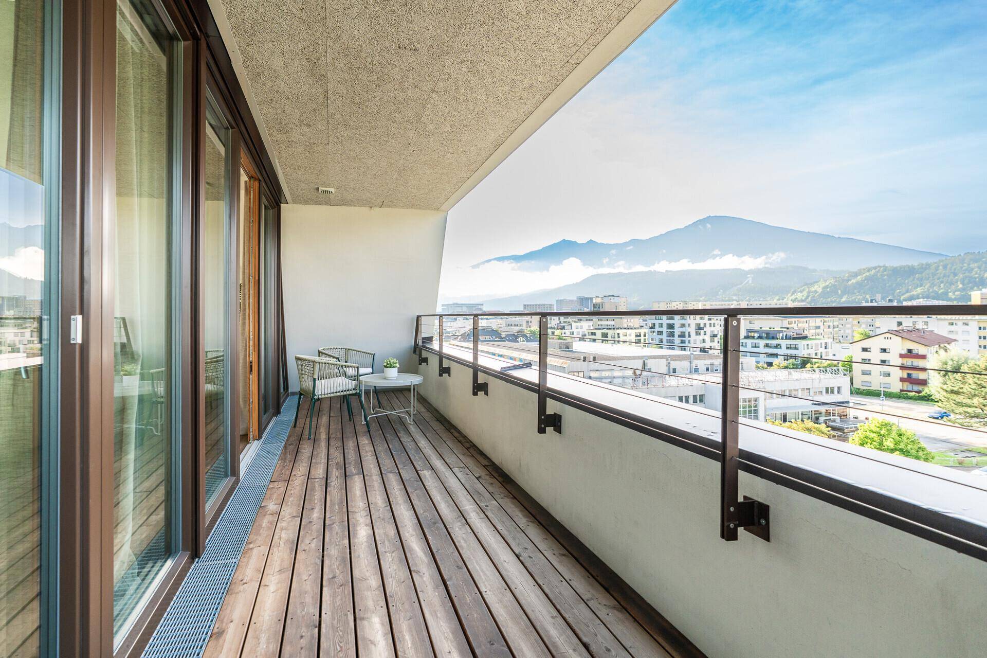 Geräumiger Balkon mit Holzbelag, Sitzgelegenheiten und weitem Blick auf Stadt und Berge.