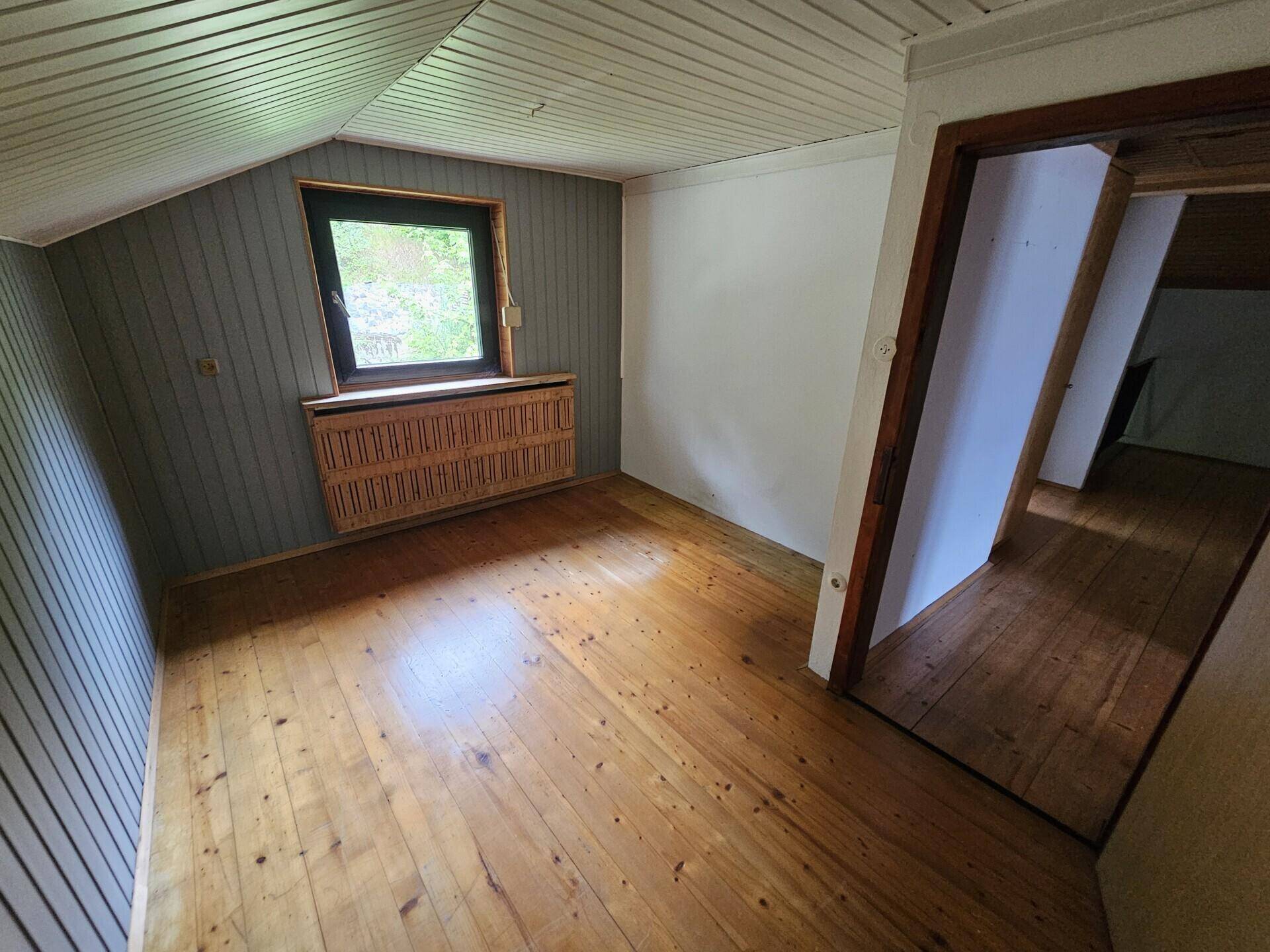 Gemütliches Dachzimmer mit Holzverkleidung, Holzboden und Fenster mit Grünblick.