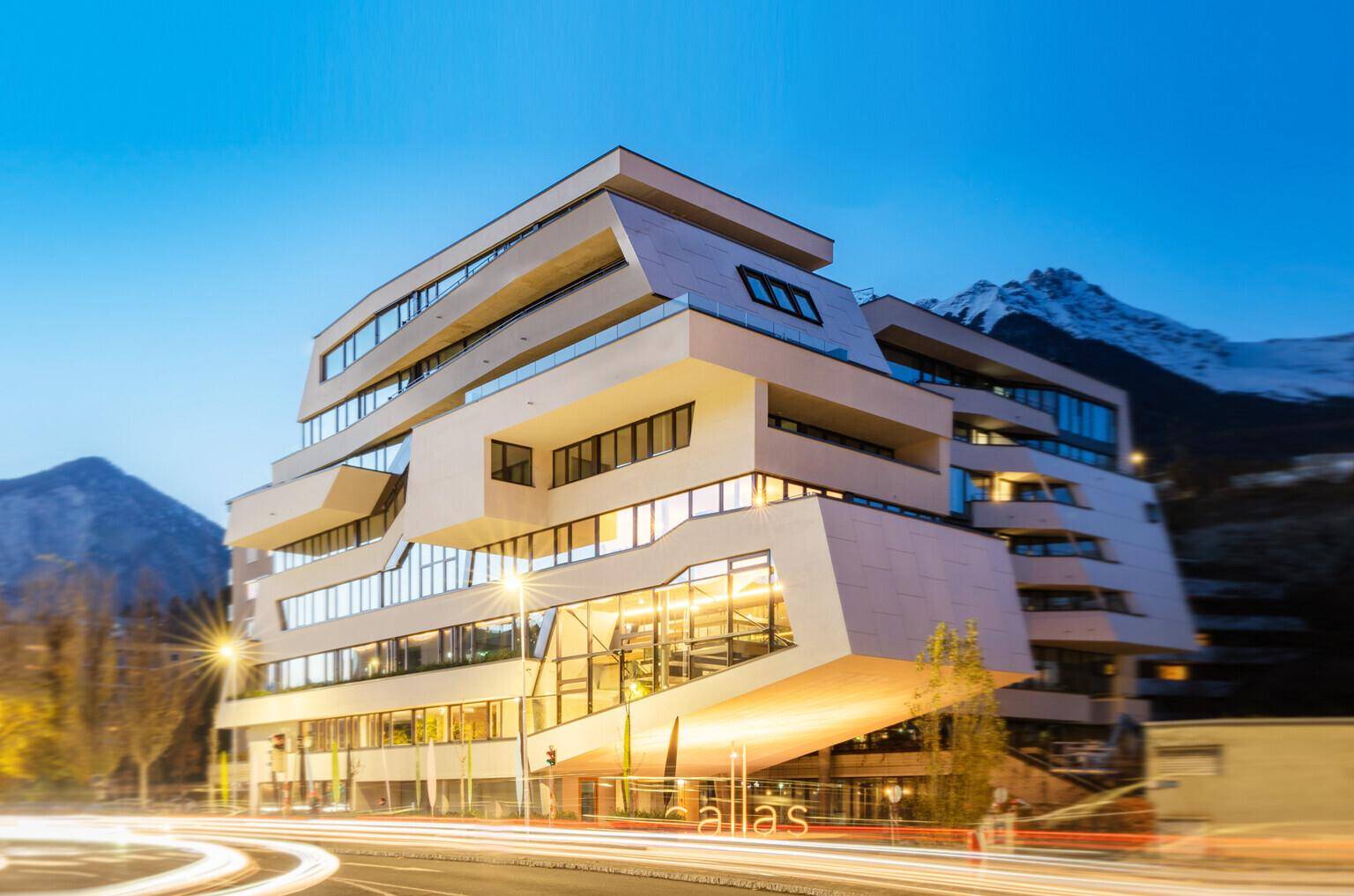 Moderne Architekturfassade eines Gebäudes bei Dämmerung mit beleuchteten Fenstern und Bergblick.