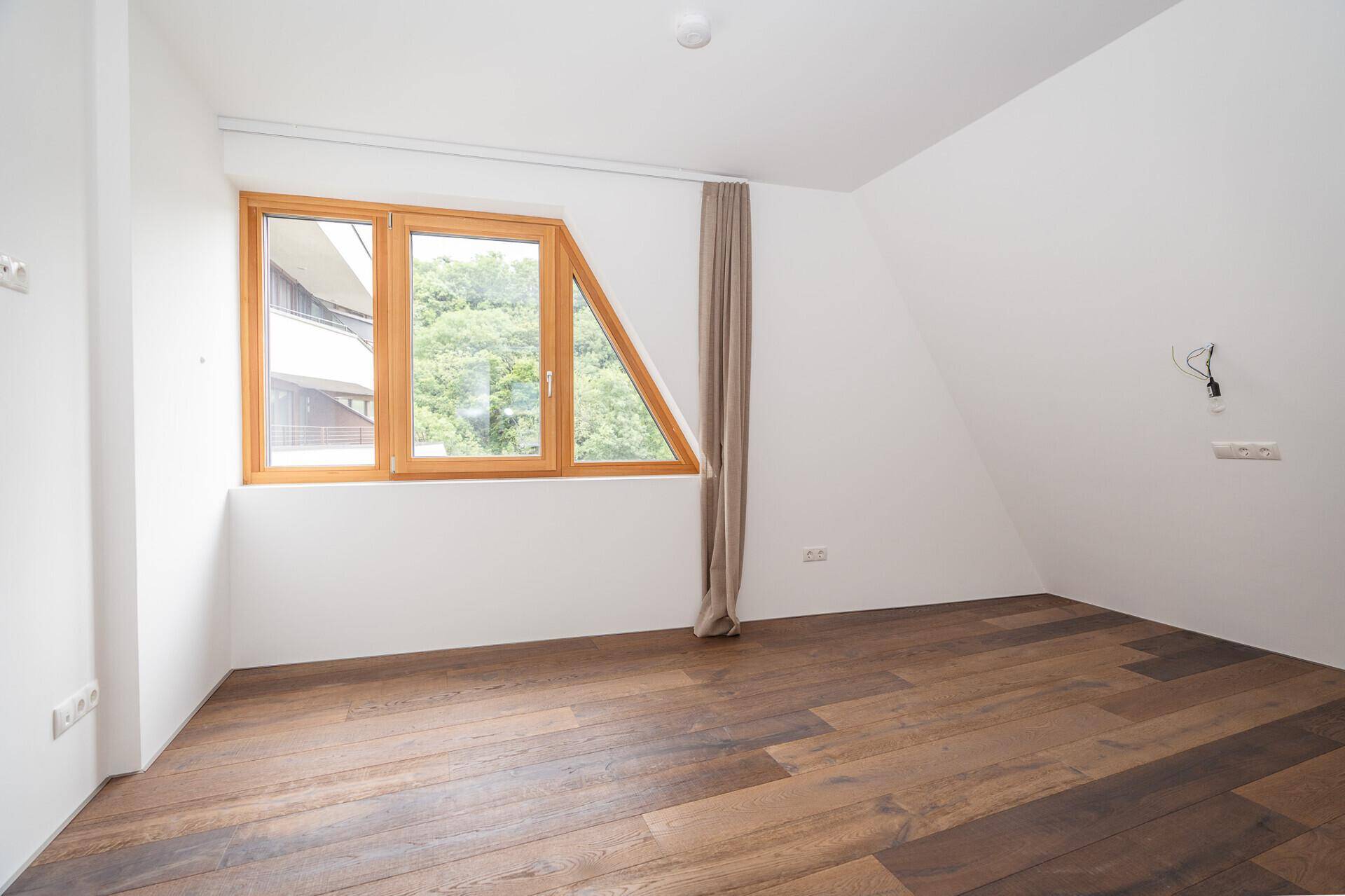 Helles Zimmer mit Holzboden, Dachschräge und einem Fenster mit Blick ins Grüne.
