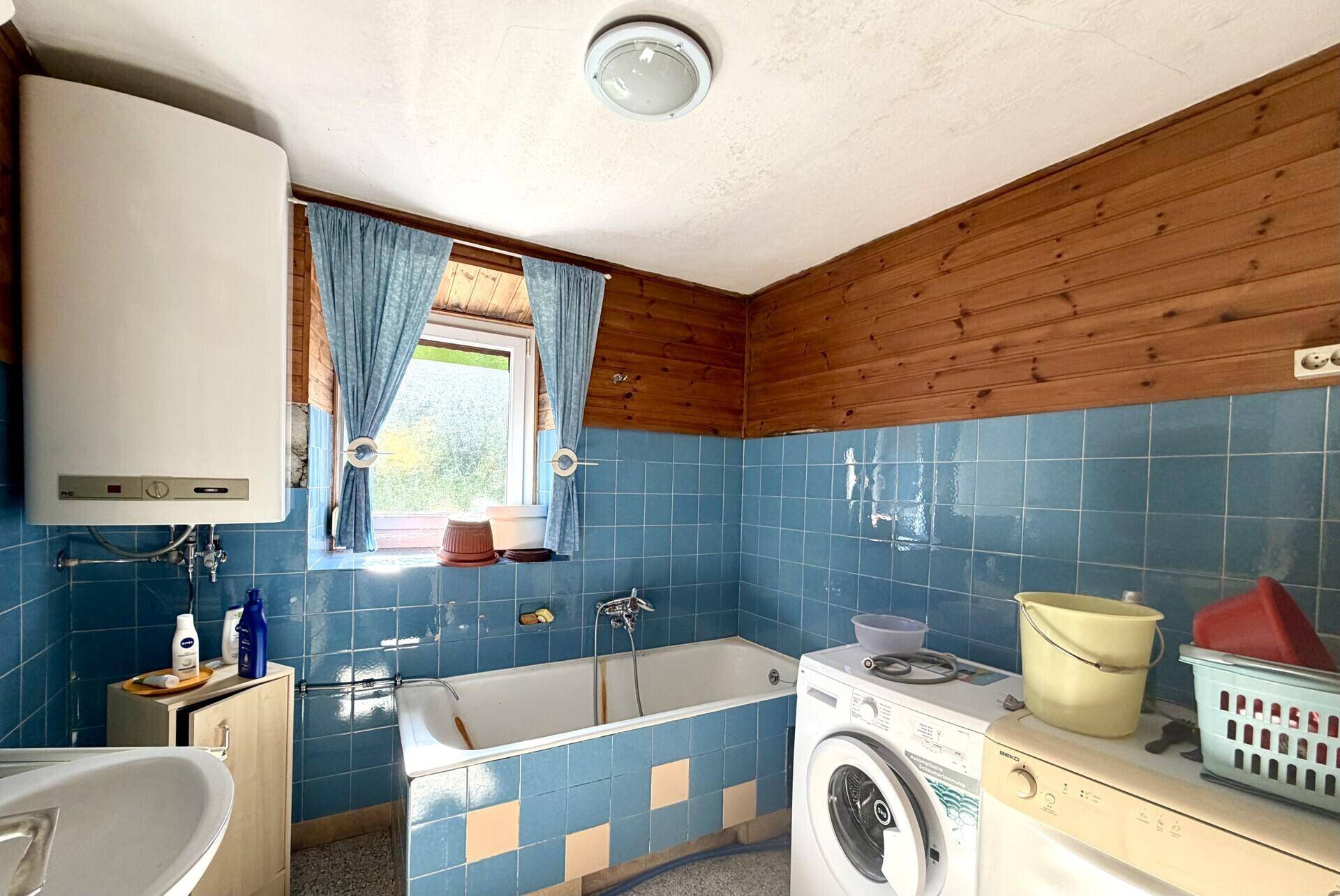 Badezimmer mit blauen und beigen Fliesen, Badewanne und Waschmaschine unter einem Fenster.