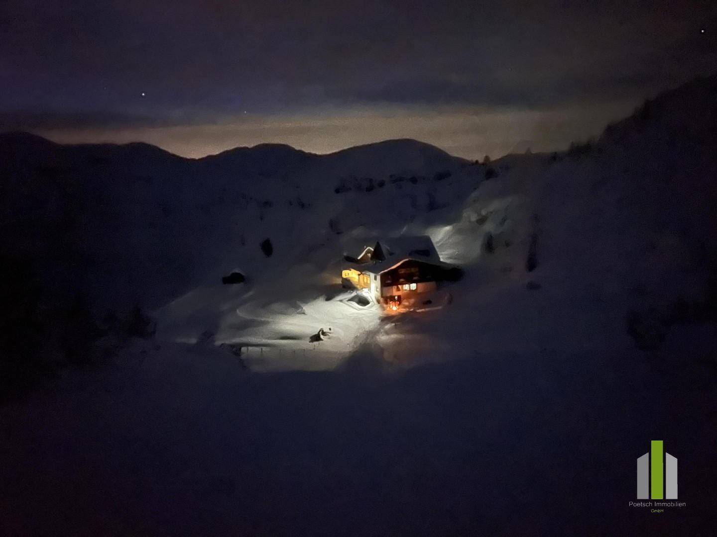 Eine Berghütte in einer verschneiten Winterlandschaft bei Nacht, beleuchtet unter einem dunklen Himmel und umgeben von Bergen.