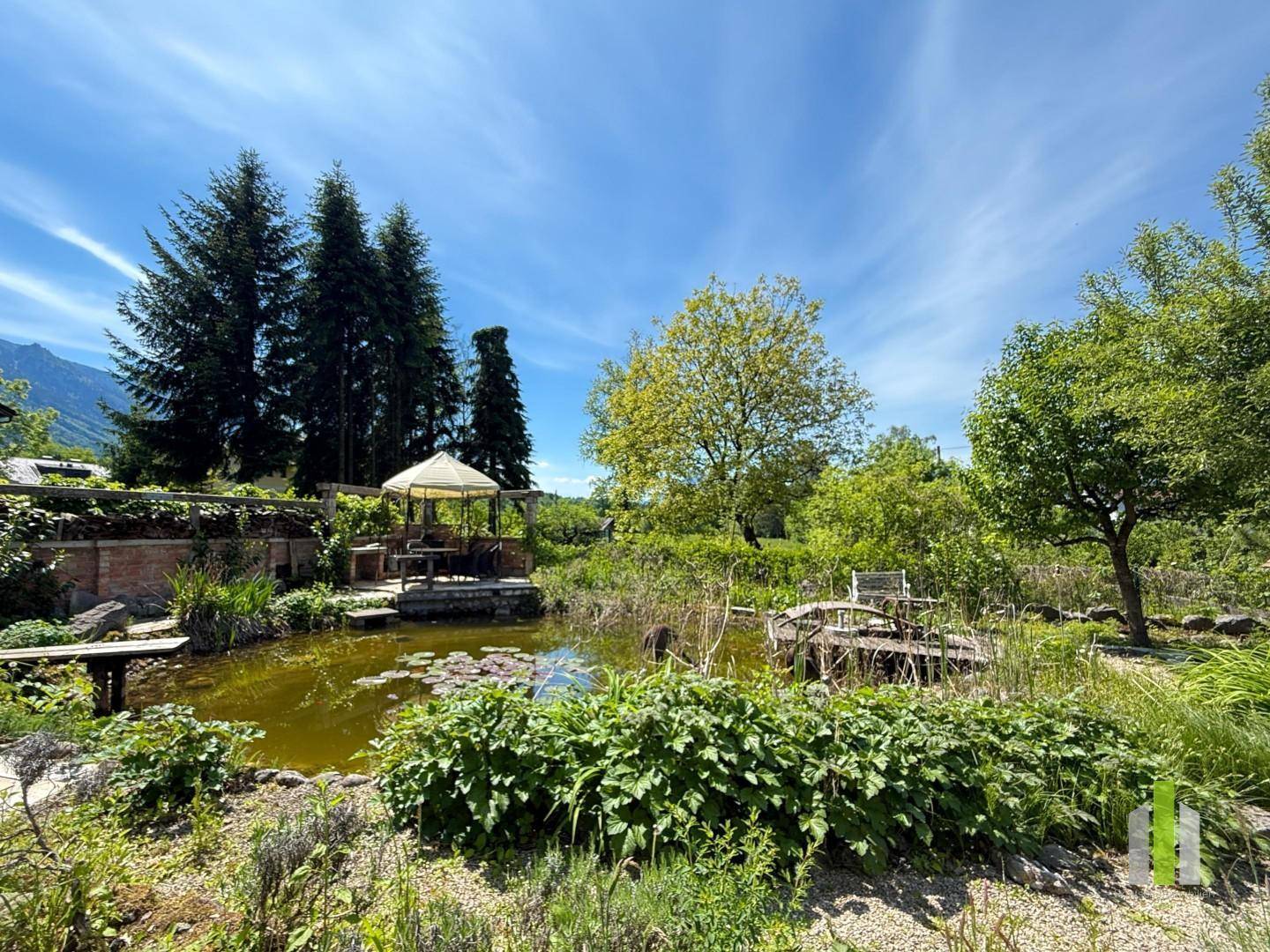 Idyllischer Garten mit einem Teich, einem Pavillon und üppiger Vegetation unter blauem Himmel.