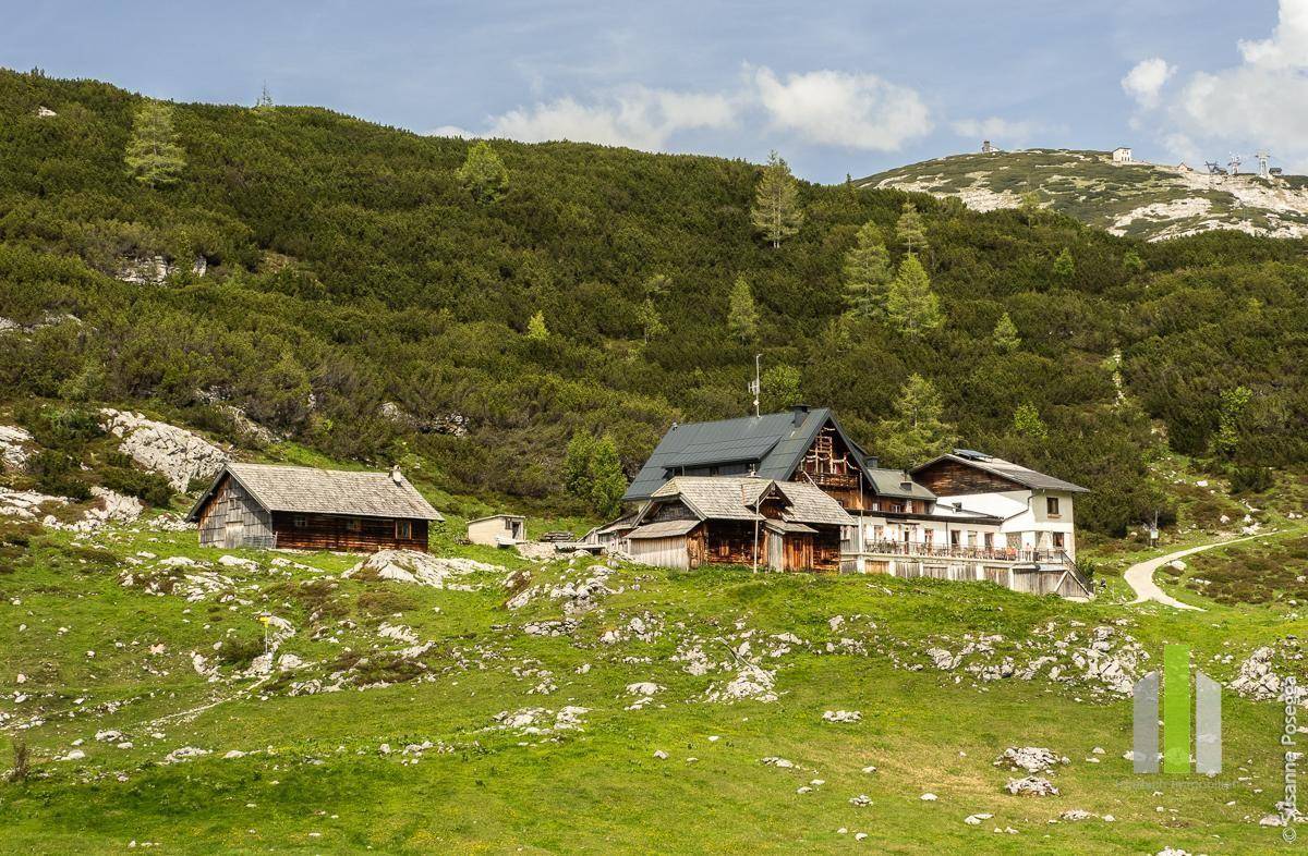 Traditionelles Gebäude in einer grünen Berglandschaft mit felsigen Hügeln und üppiger Vegetation unter blauem Himmel.