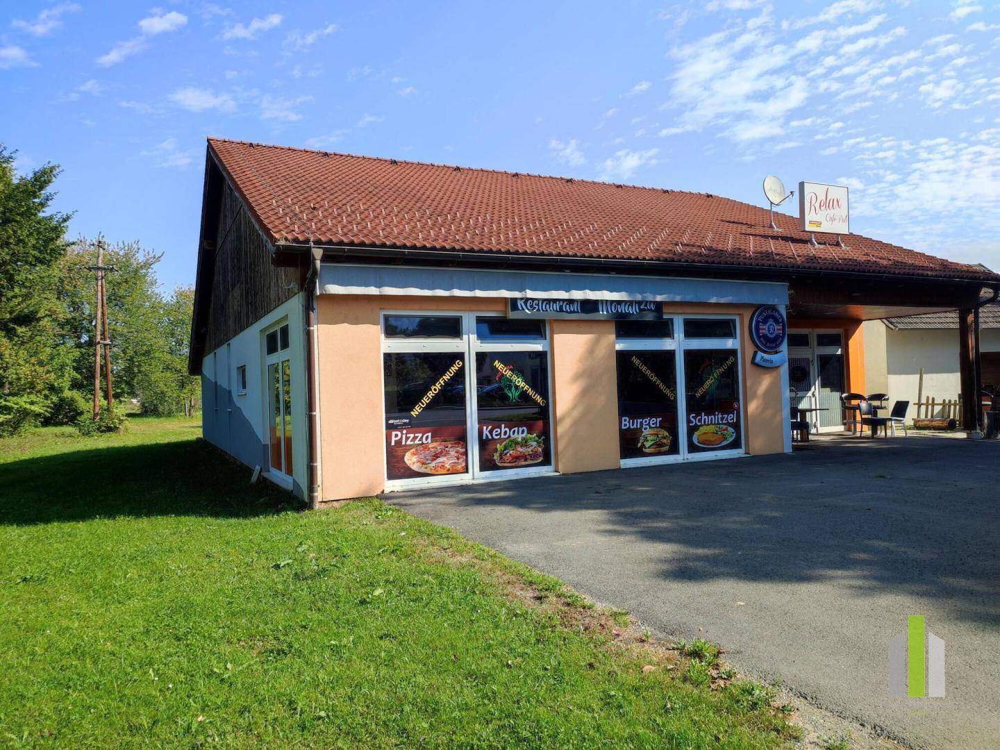 Seitenansicht eines Restaurants mit Schaufenstern, Werbeaufklebern und einem angrenzenden Grünbereich.