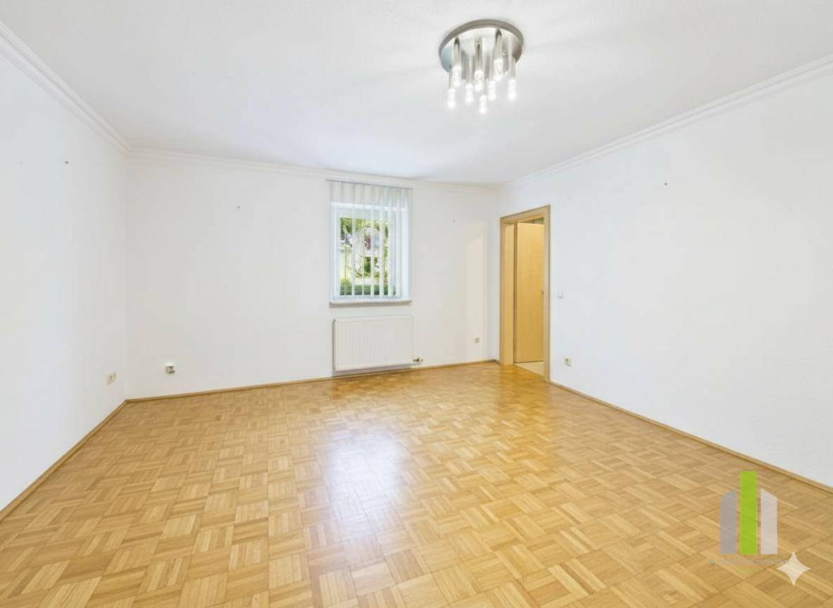 Helles Zimmer mit Parkettboden, einem Fenster und einem Heizkörper unter dem Fenster.