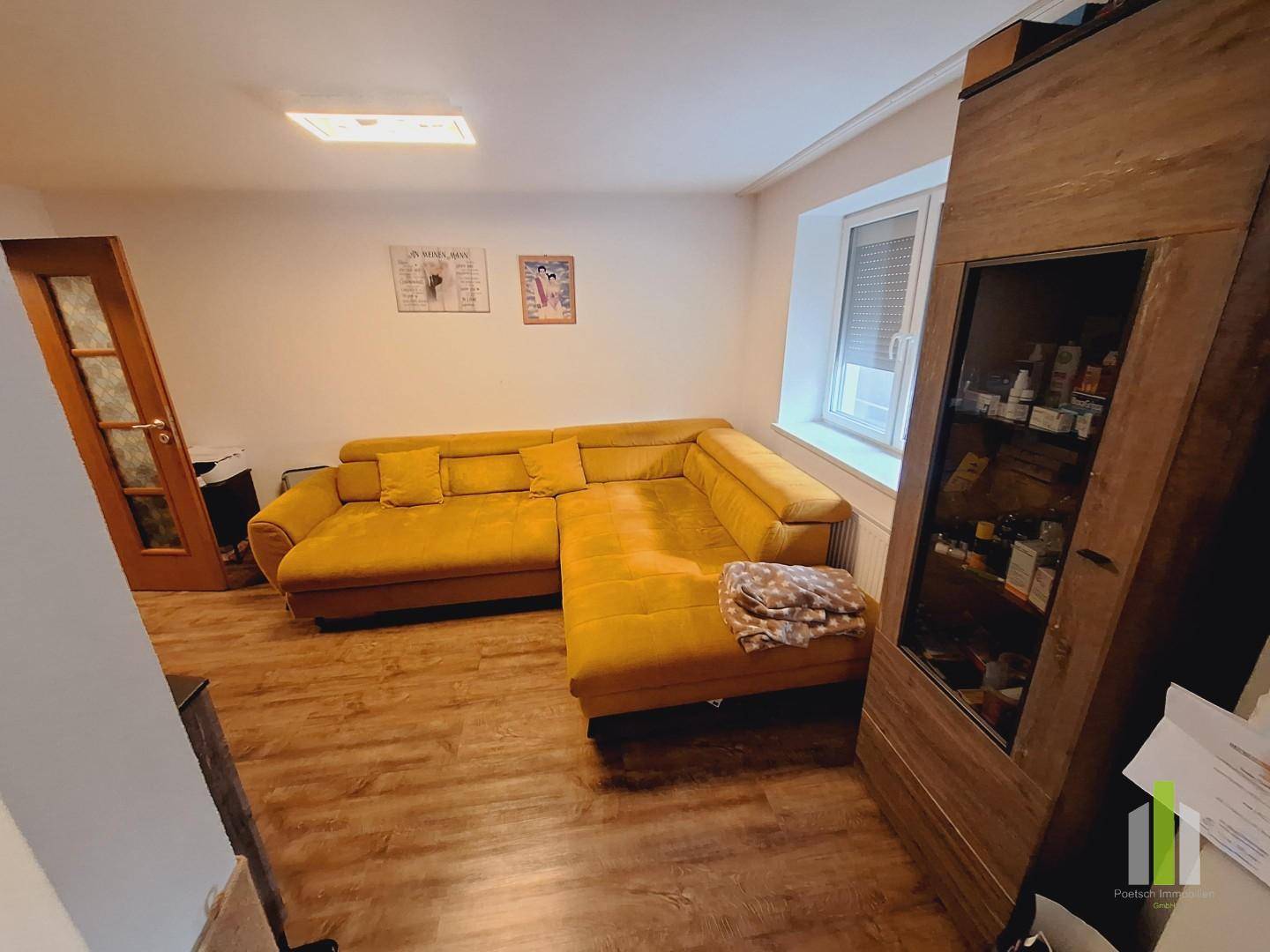 Gemütliches Wohnzimmer mit einem großen gelben Ecksofa und Laminatboden, hell durch das Fenster.