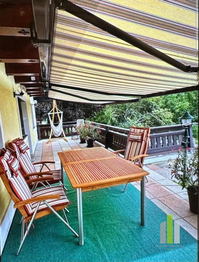 Geräumige Terrasse mit Markise, Holztisch und Stühlen, umgeben von üppigem Grün.