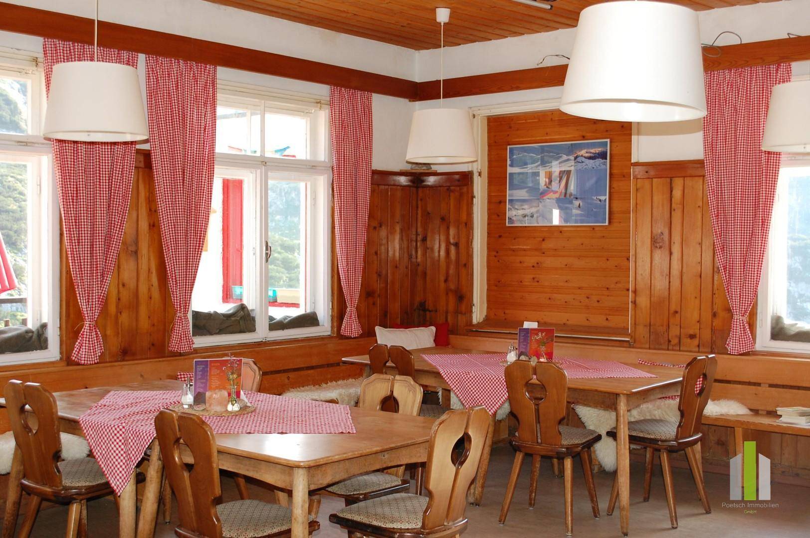 Rustikaler Essbereich mit Holztischen, Stühlen und karierten Vorhängen, umgeben von großen Fenstern.