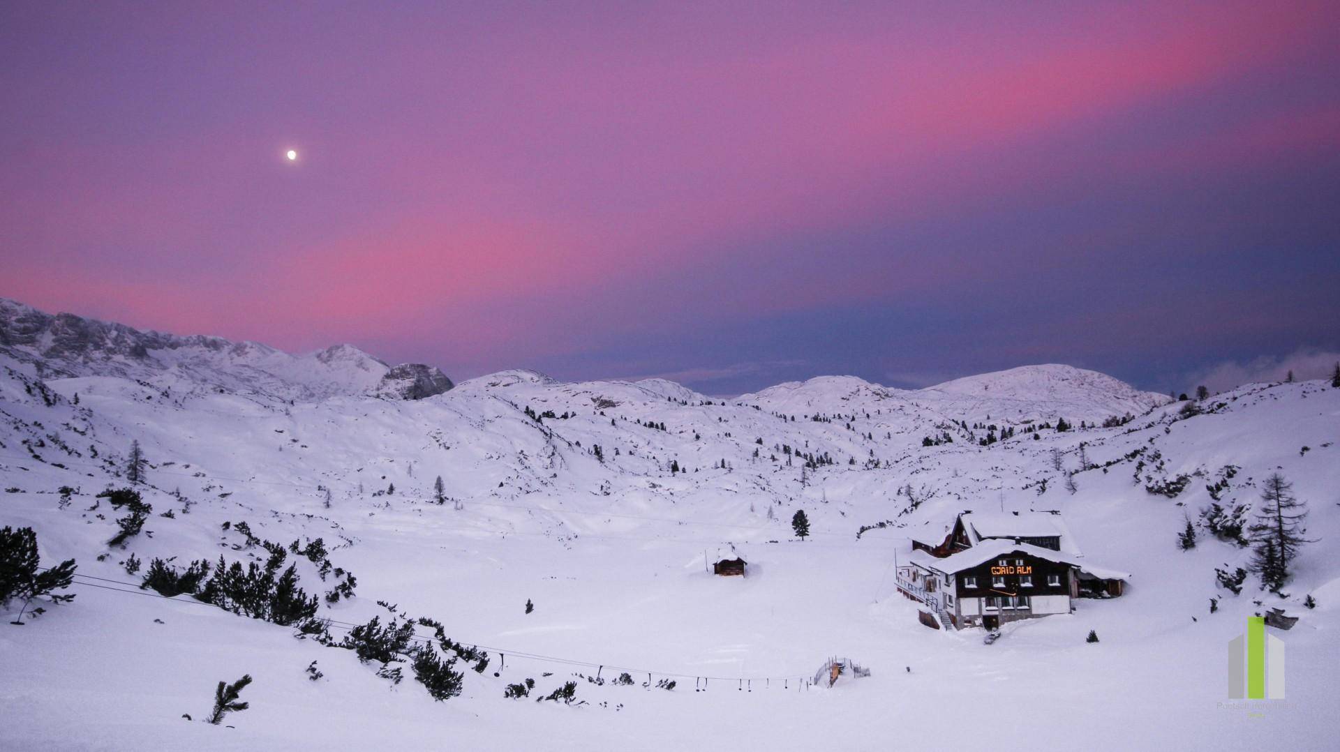 Berghütte in verschneiter Landschaft bei Sonnenuntergang mit rosa Himmel und Mond am Horizont.