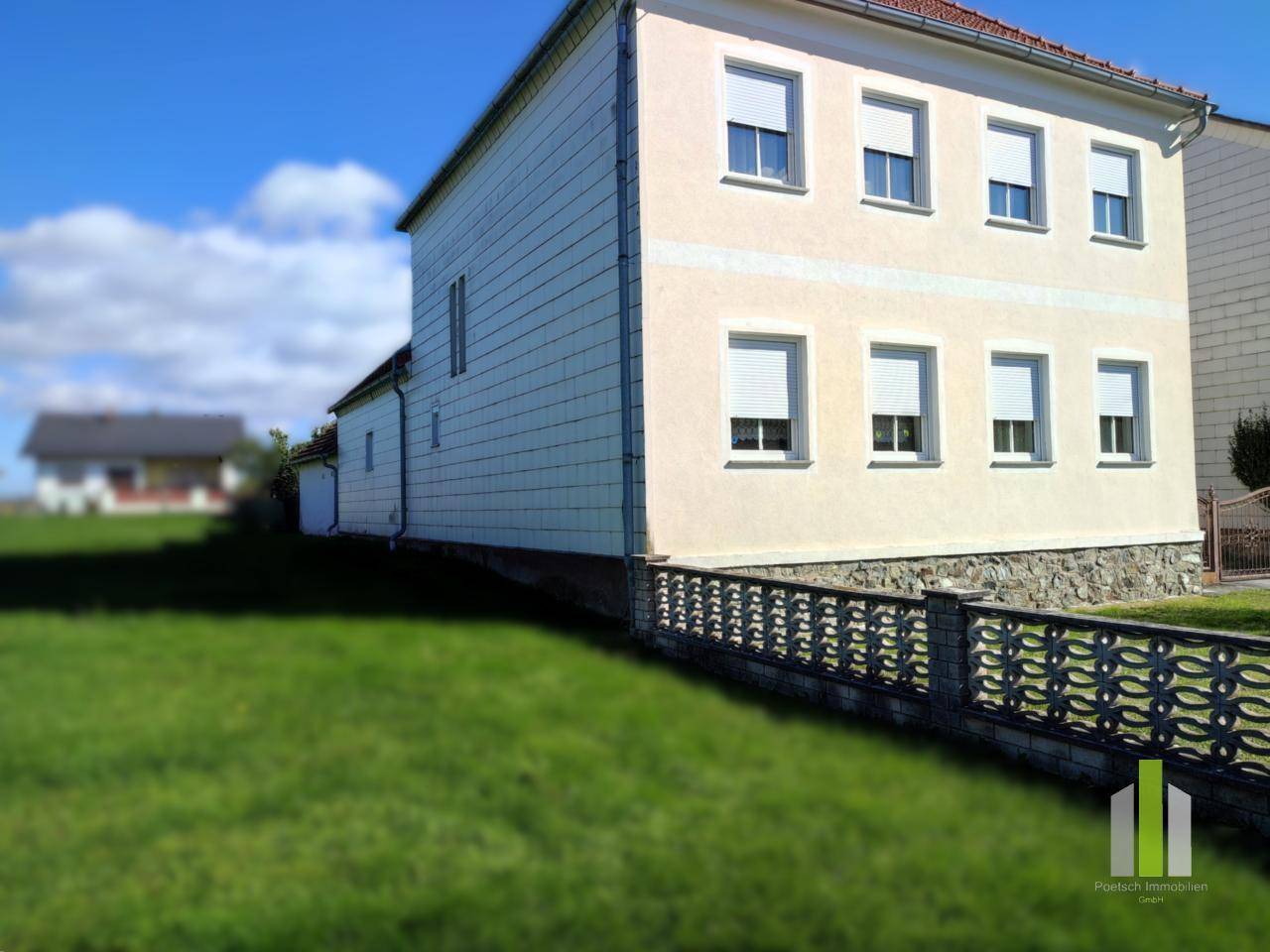 Seitenansicht des Hauses mit Rollläden, umzäuntem Gartenbereich und ländlicher Umgebung.