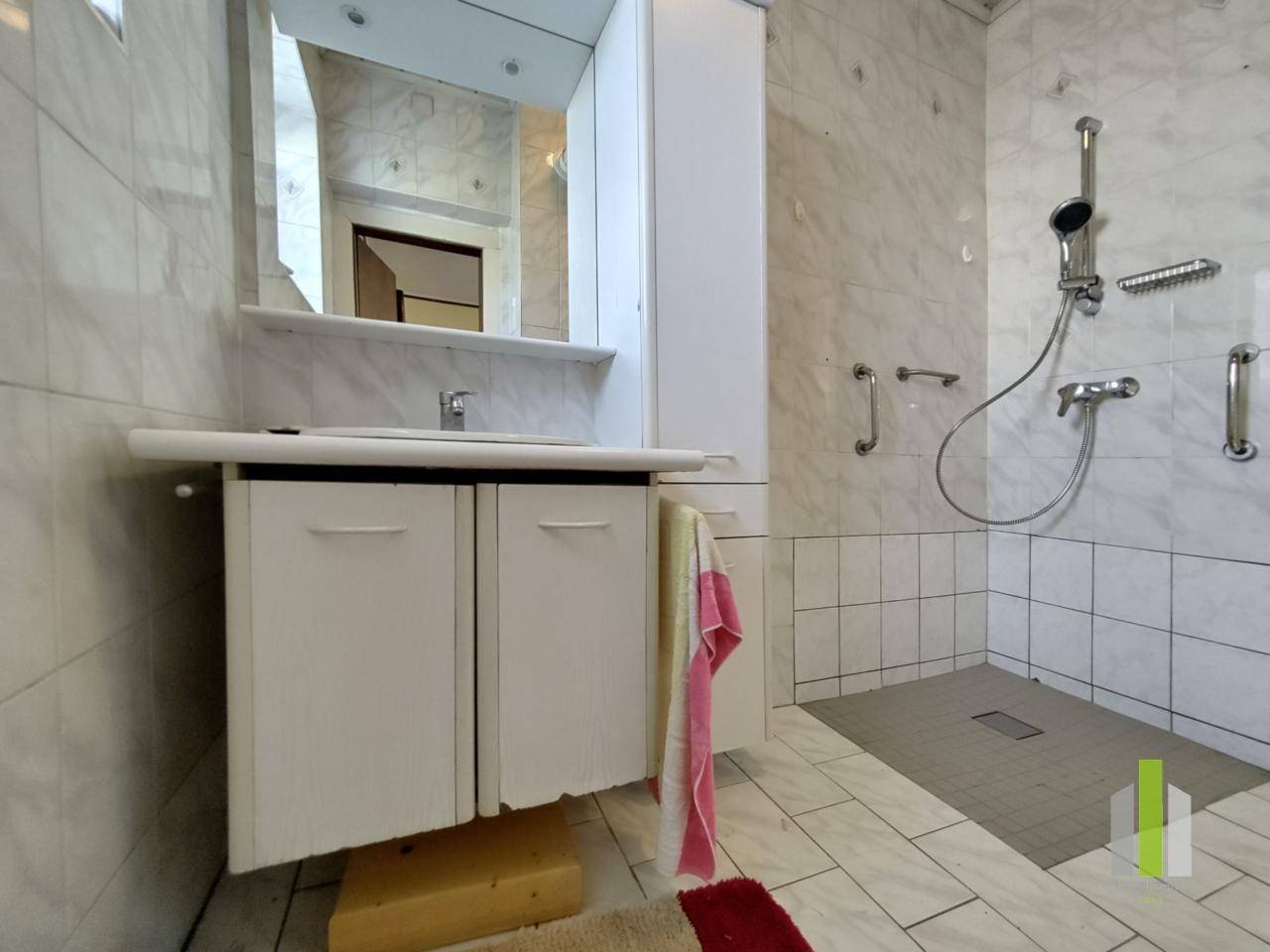 Modernisiertes Badezimmer mit weißen Marmorfliesen, ebenerdiger Dusche und Haltegriffen.