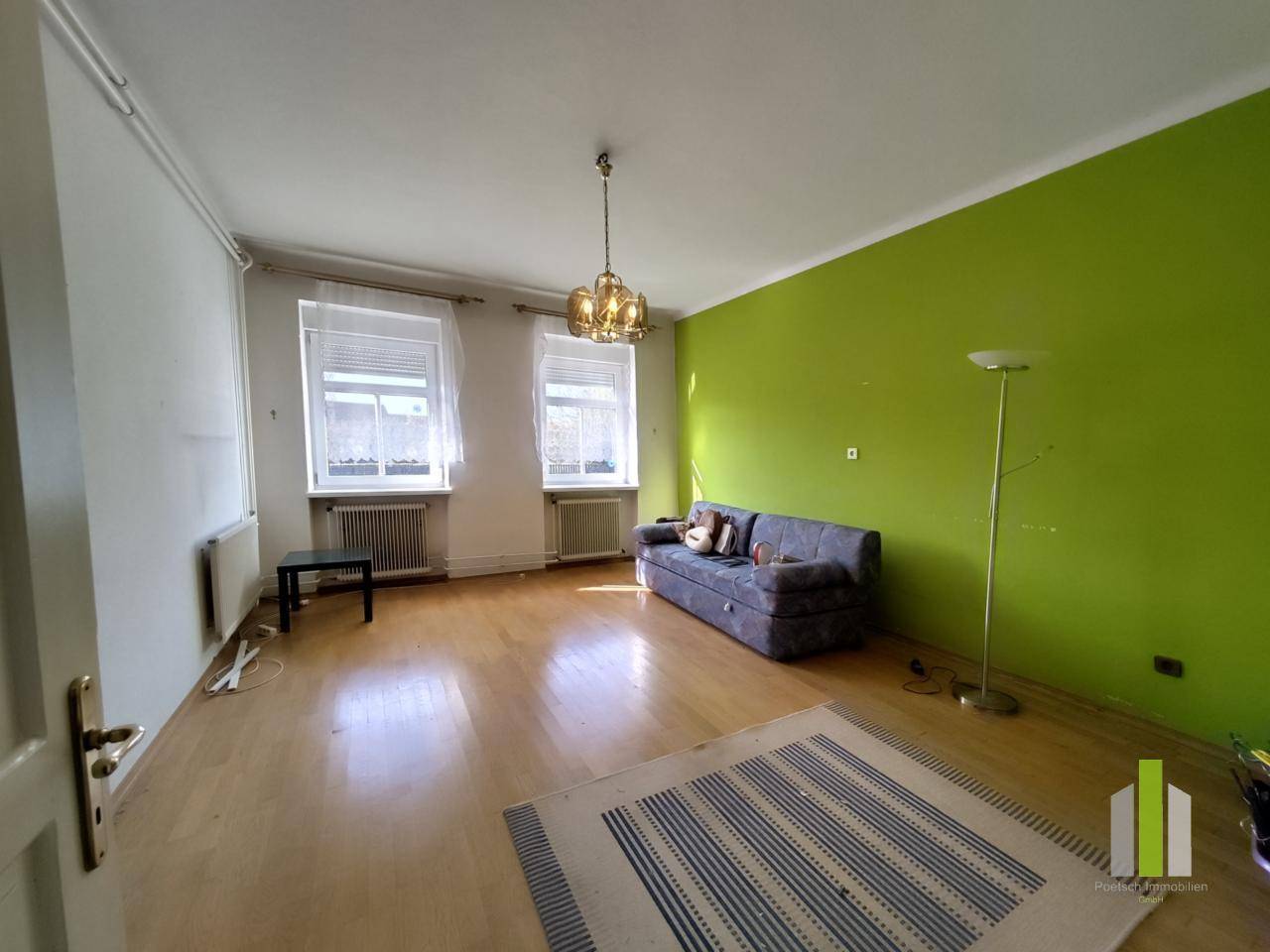 Helles Wohnzimmer mit grün gestrichener Wand, Parkettboden und zwei großen Fenstern.