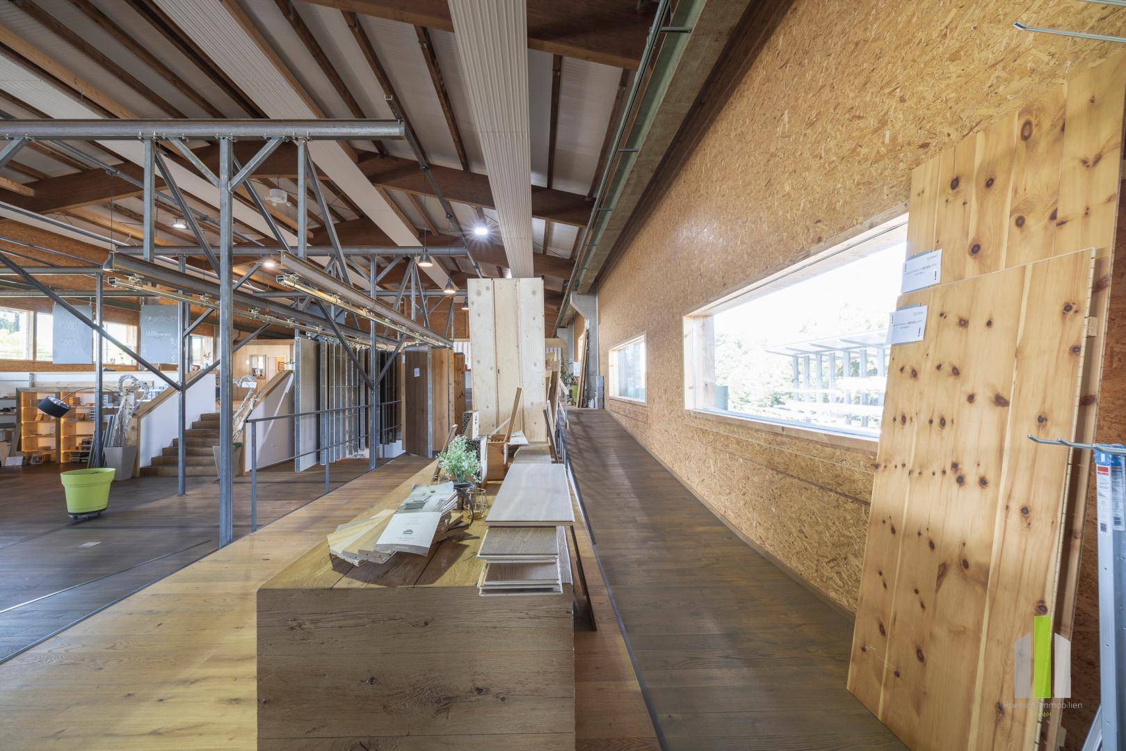 Innenansicht eines hellen Ausstellungsraums mit Holzböden, großen Fenstern und verschiedenen Holzmaterialien.