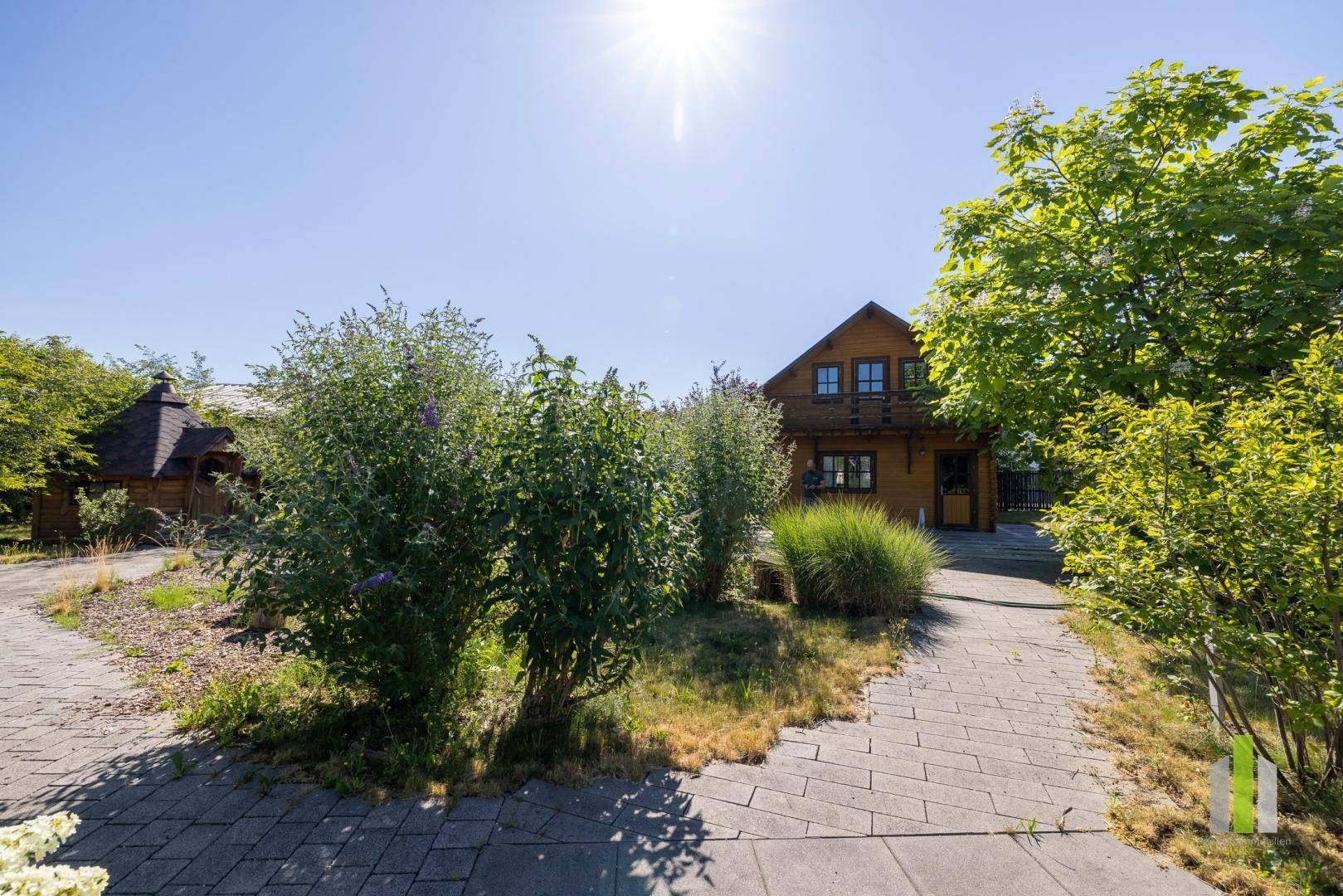 Charmantes Holzhaus mit gepflastertem Weg und üppiger Bepflanzung unter strahlend blauem Himmel.