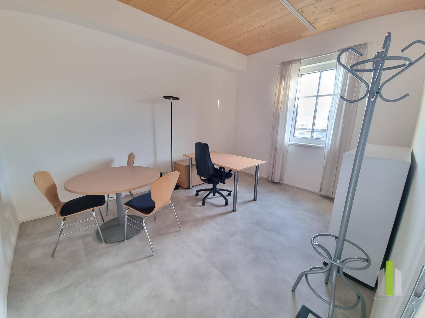 Kleines Büro mit Schreibtisch, Besprechungstisch und Fenster, ausgestattet mit Fliesenboden und Holzbalkendecke.