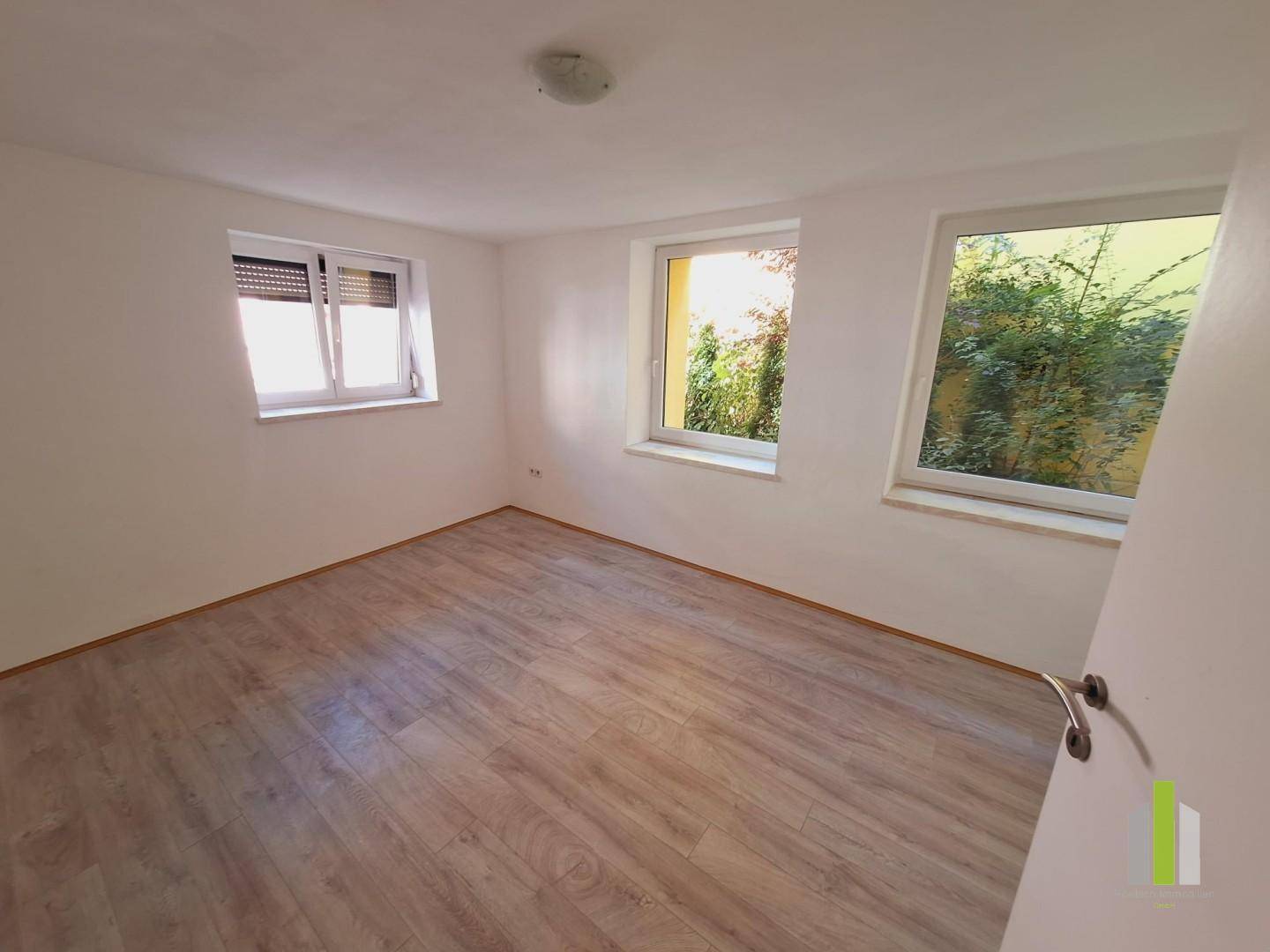 Leeres Zimmer mit hellem Laminatboden und zwei Fenstern, eines davon mit Rollladen.
