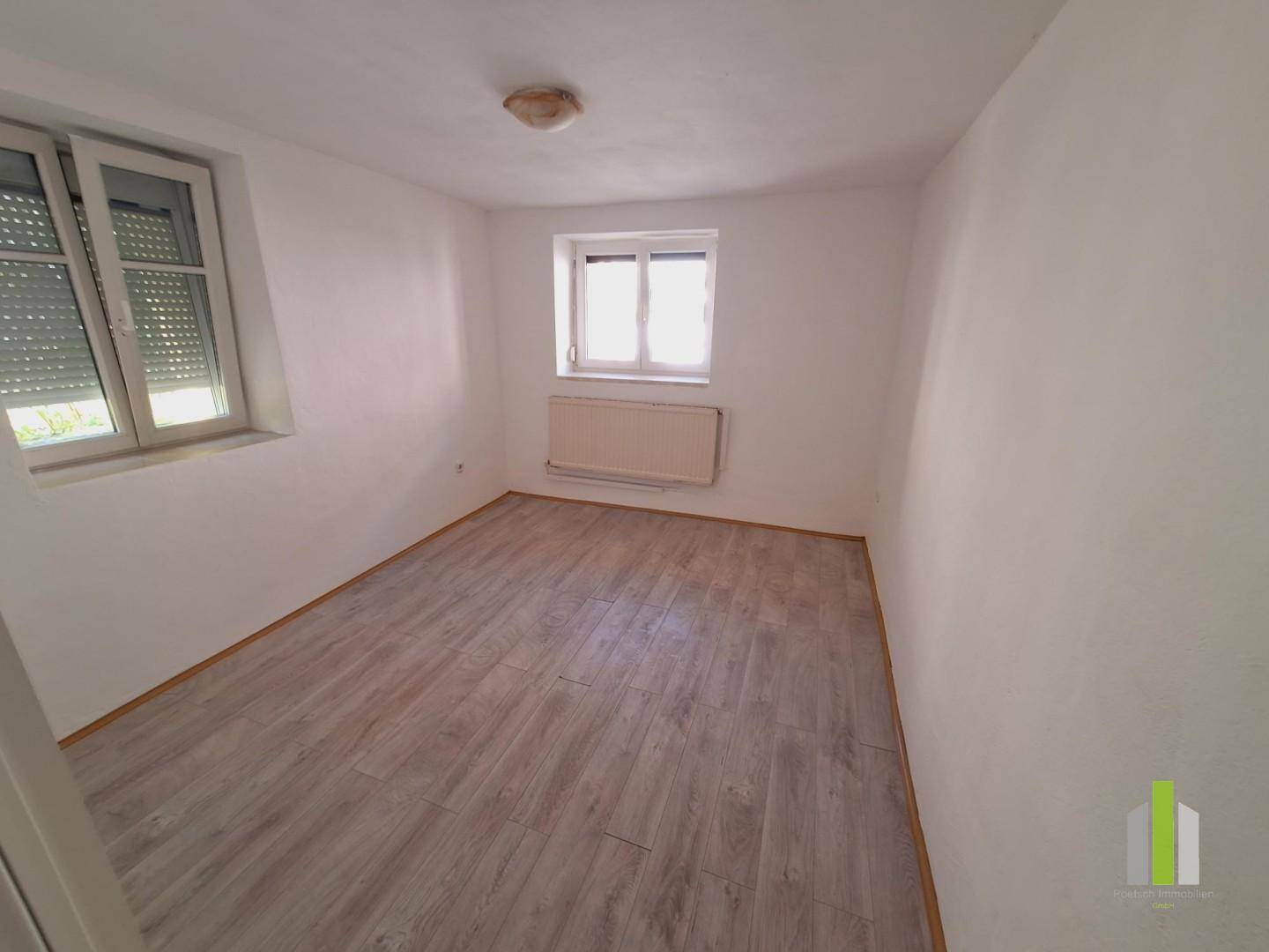 Helles Zimmer mit Laminatboden, zwei Fenstern und Heizkörper unter einem Fenster.