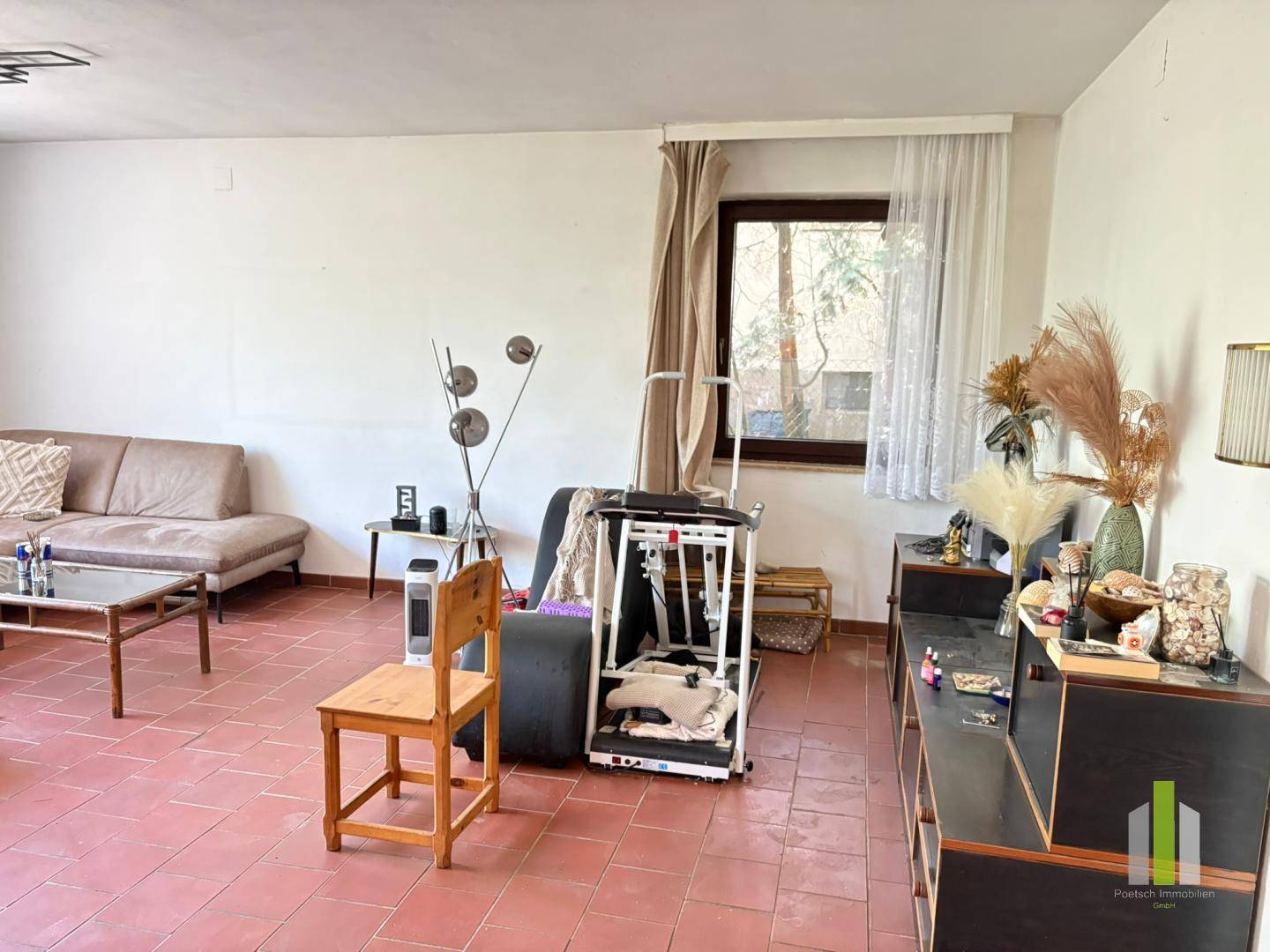 Geräumiges Wohnzimmer mit Terrakottafliesen, Sofa und einem Fenster, das viel Tageslicht hereinlässt.