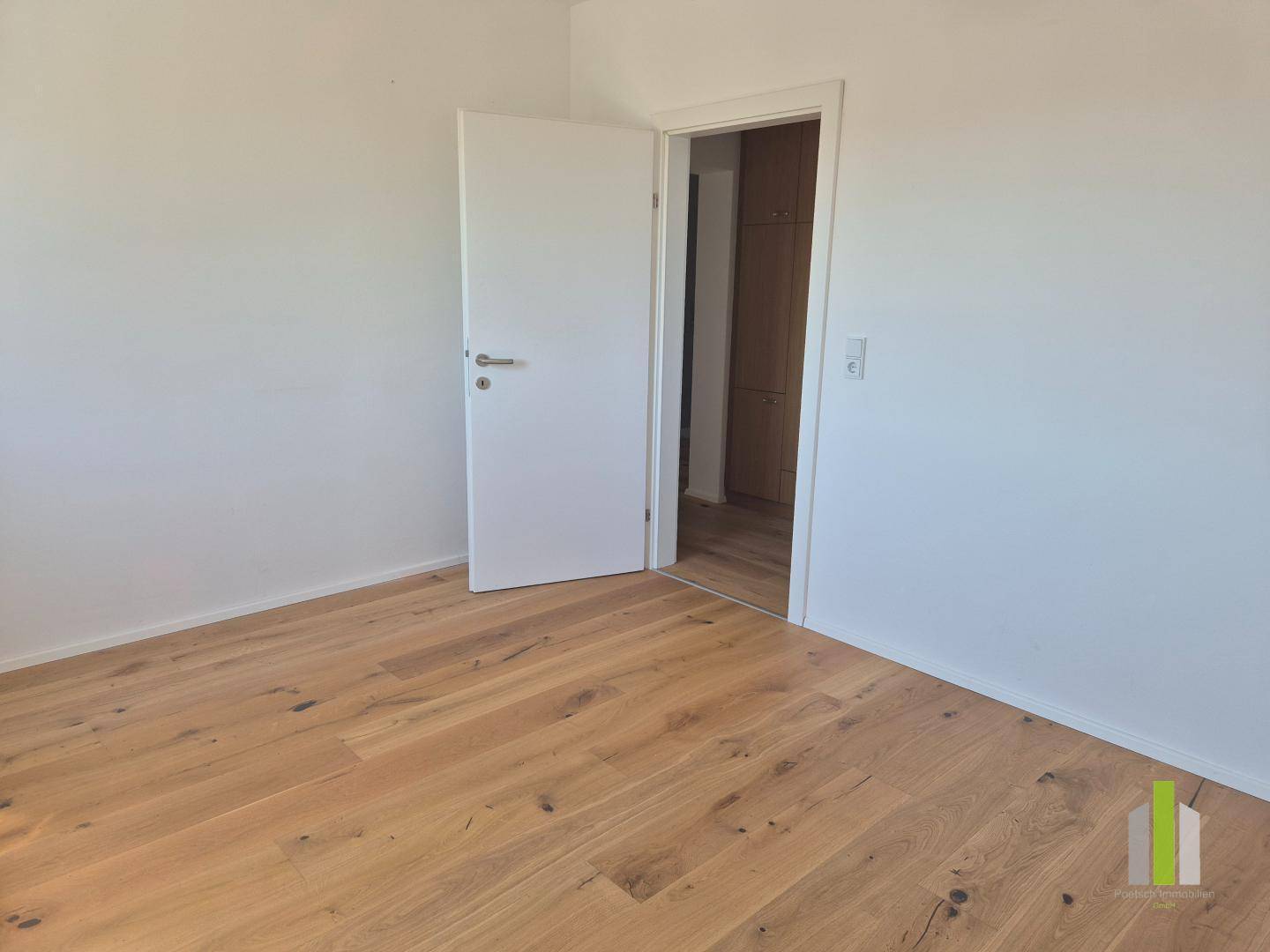 Geräumiges Zimmer mit Holzboden und weißer Tür, die zu einem angrenzenden Bereich führt.