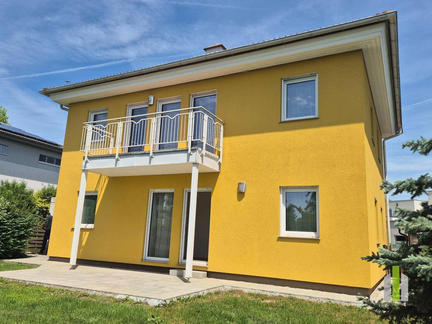 Rückseite des Hauses mit gelber Fassade, Balkon und einer gepflasterten Terrasse im Garten.