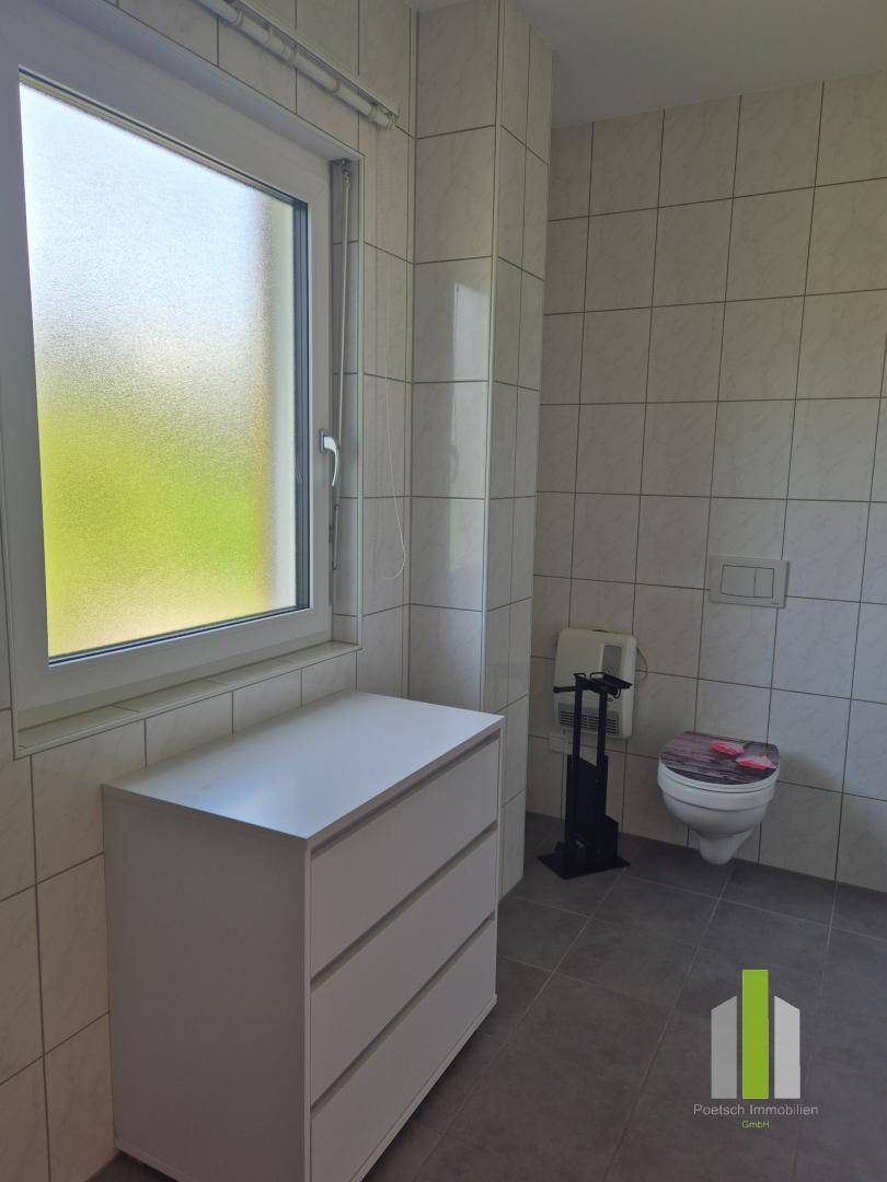Badezimmer mit Toilette, Fenster und Kommode, hell gefliest und mit natürlicher Belüftung.