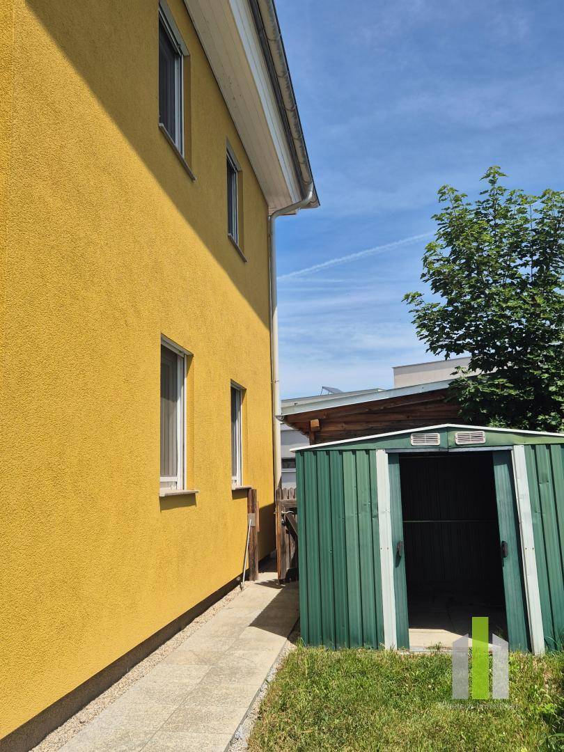 Seitenansicht des Hauses mit gelber Fassade und einem grünen Gartenschuppen auf dem Rasen.