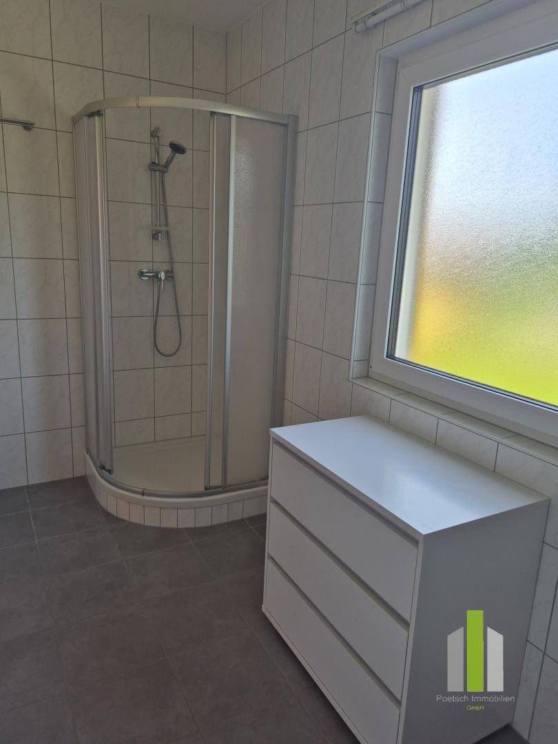 Badezimmer mit Eckduschkabine, Fenster und Kommode, hell gefliest und mit Tageslicht.