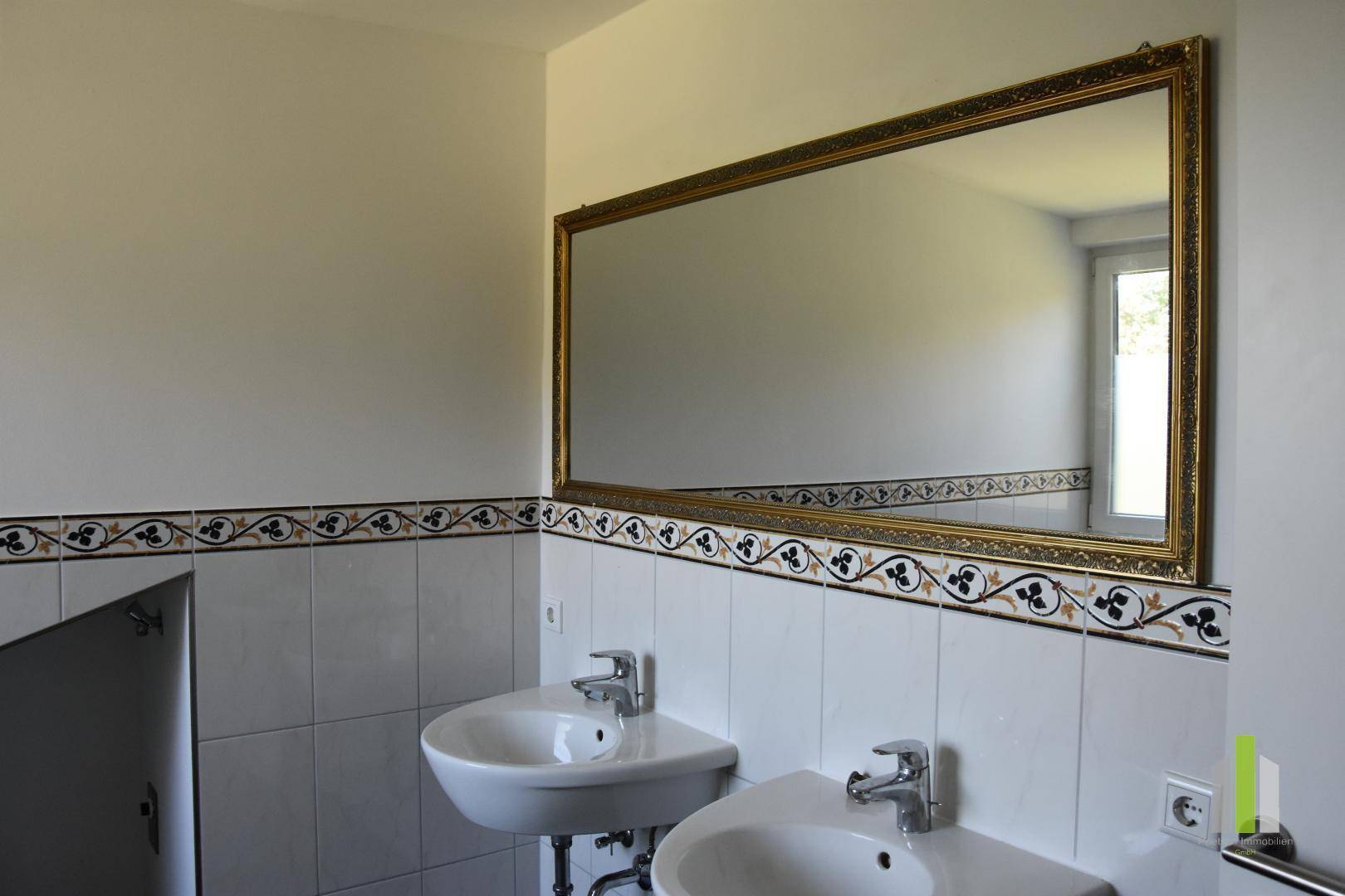 Badezimmer mit Doppelwaschbecken, großem Spiegel und dekorativer Fliesenbordüre.