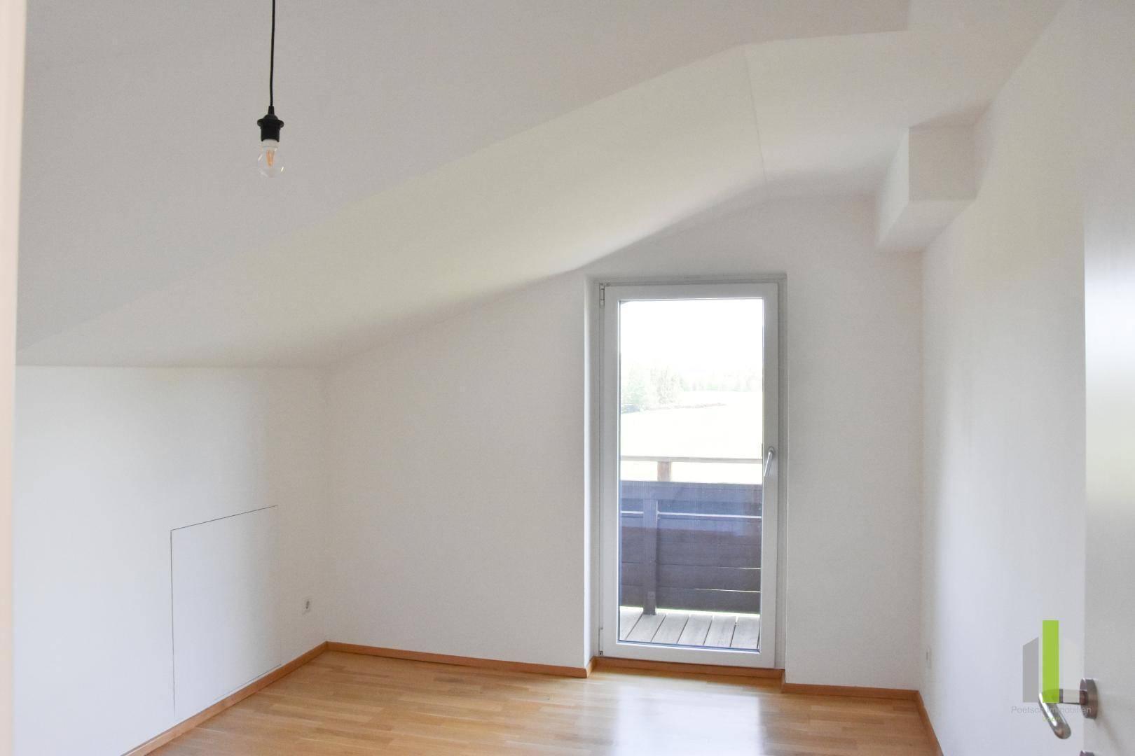 Helles Zimmer mit Holzboden, Dachschrägen und Zugang zu einem kleinen Balkon.
