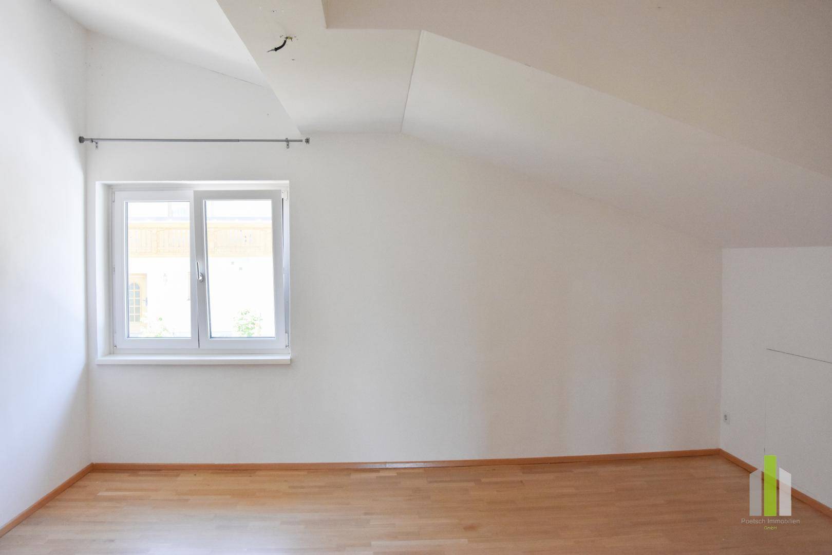Geräumiges Zimmer mit Holzboden, Dachschrägen und einem Fenster mit Blick nach draußen.