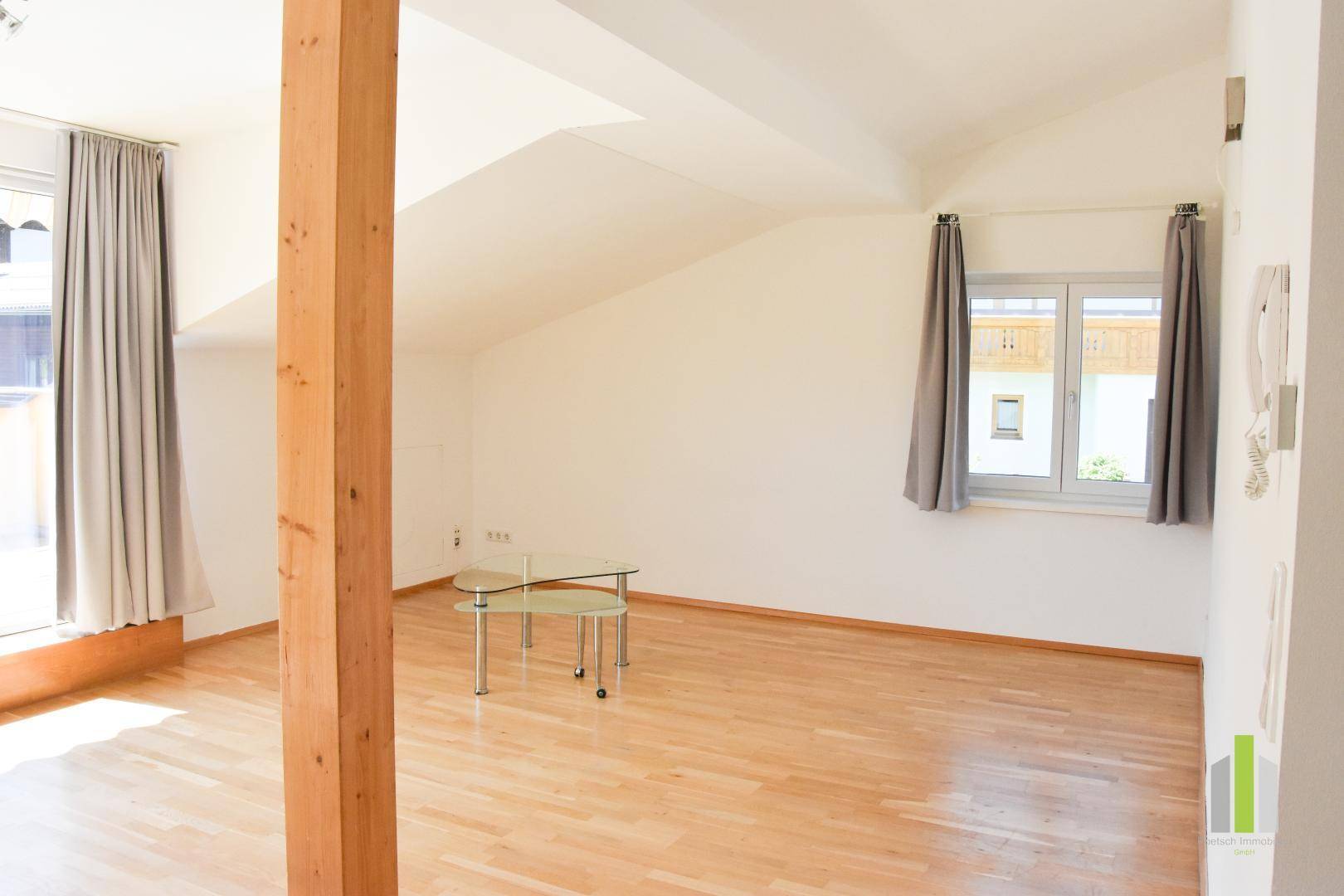 Heller Wohnbereich mit Holzboden, Dachschrägen und zwei Fenstern mit Blick ins Grüne.