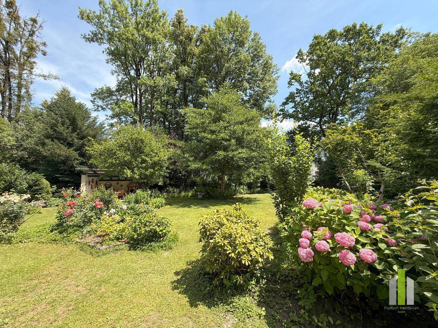 Gepflegter, weitläufiger Garten mit Rasenflächen, blühenden Sträuchern und hohen Bäumen im Hintergrund.