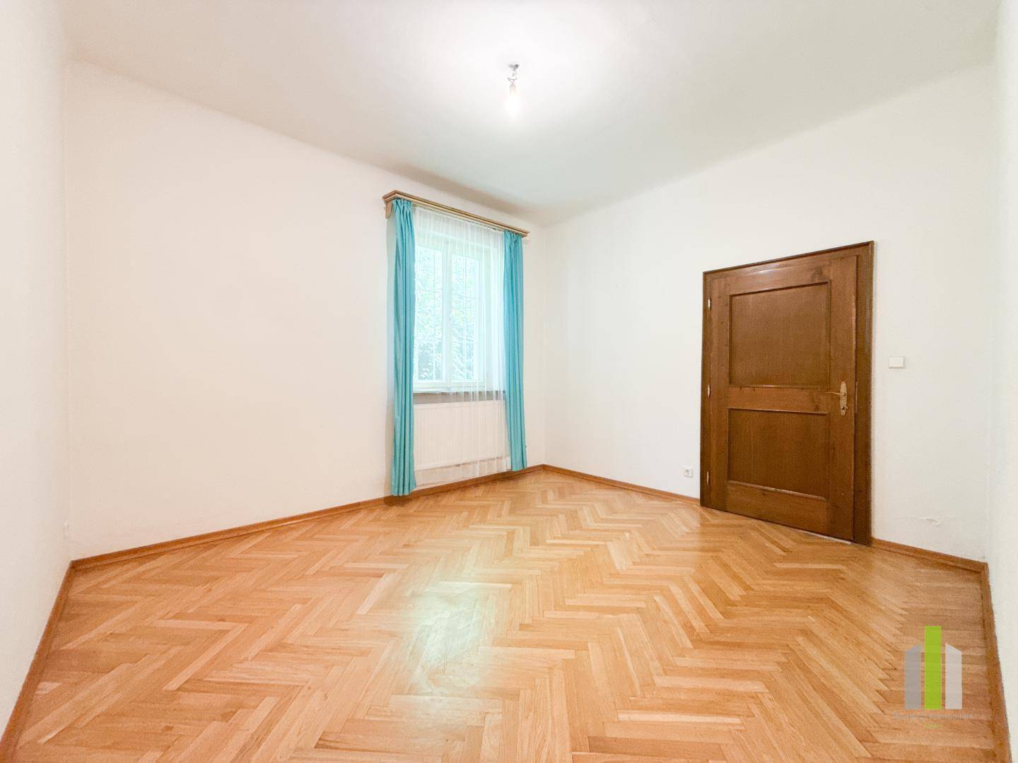 Leeres Schlafzimmer mit Fischgrätparkettboden und einem Fenster mit blauen Vorhängen.