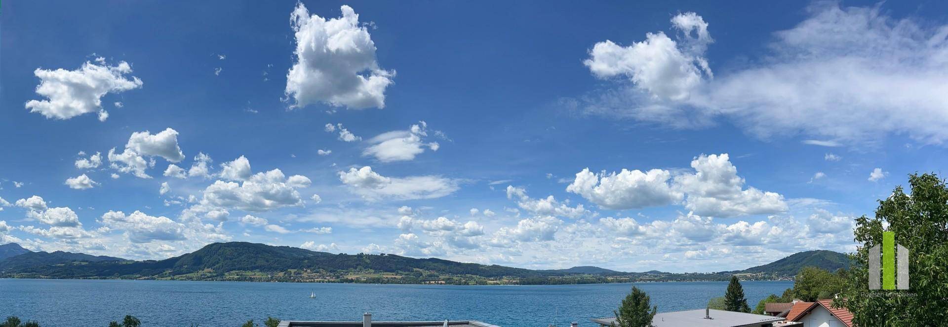 Panoramaaufnahme eines weiten Sees umgeben von Bergen unter einem strahlend blauen Himmel mit vielen Wolken.