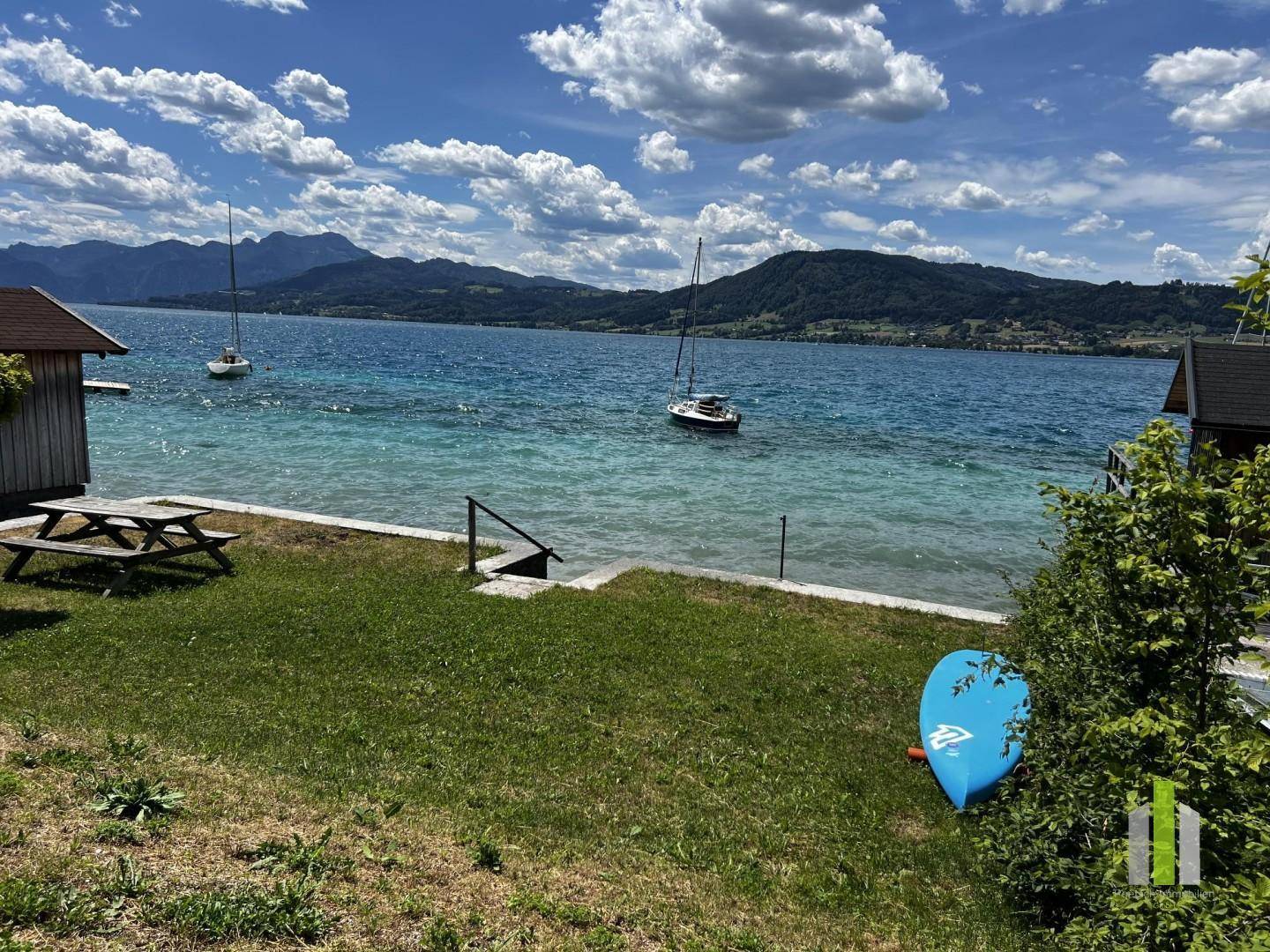 Idyllischer Gartenbereich am Seeufer mit Picknicktisch, Segelbooten und malerischem Bergblick.