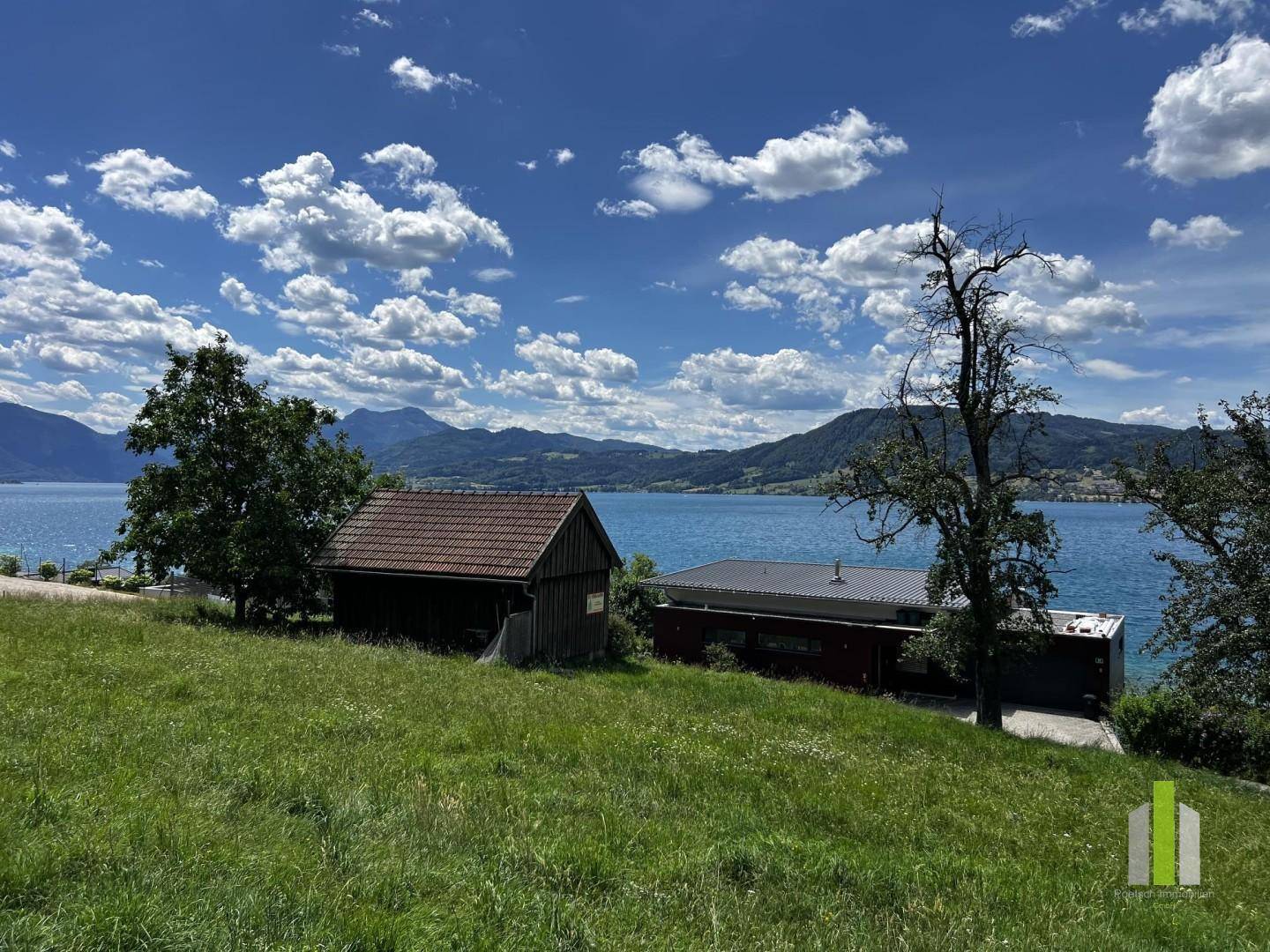 Malerischer Blick auf den See mit Bergen, grüner Wiese und kleinen Gebäuden am Ufer unter blauem Himmel.