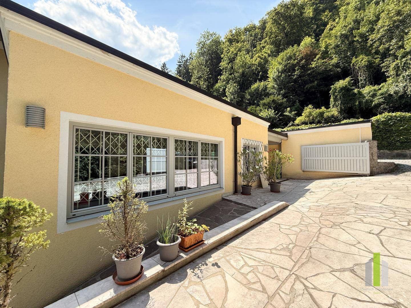 Gelbe Hausfassade mit gepflastertem Bereich und umgebender Natur unter blauem Himmel.