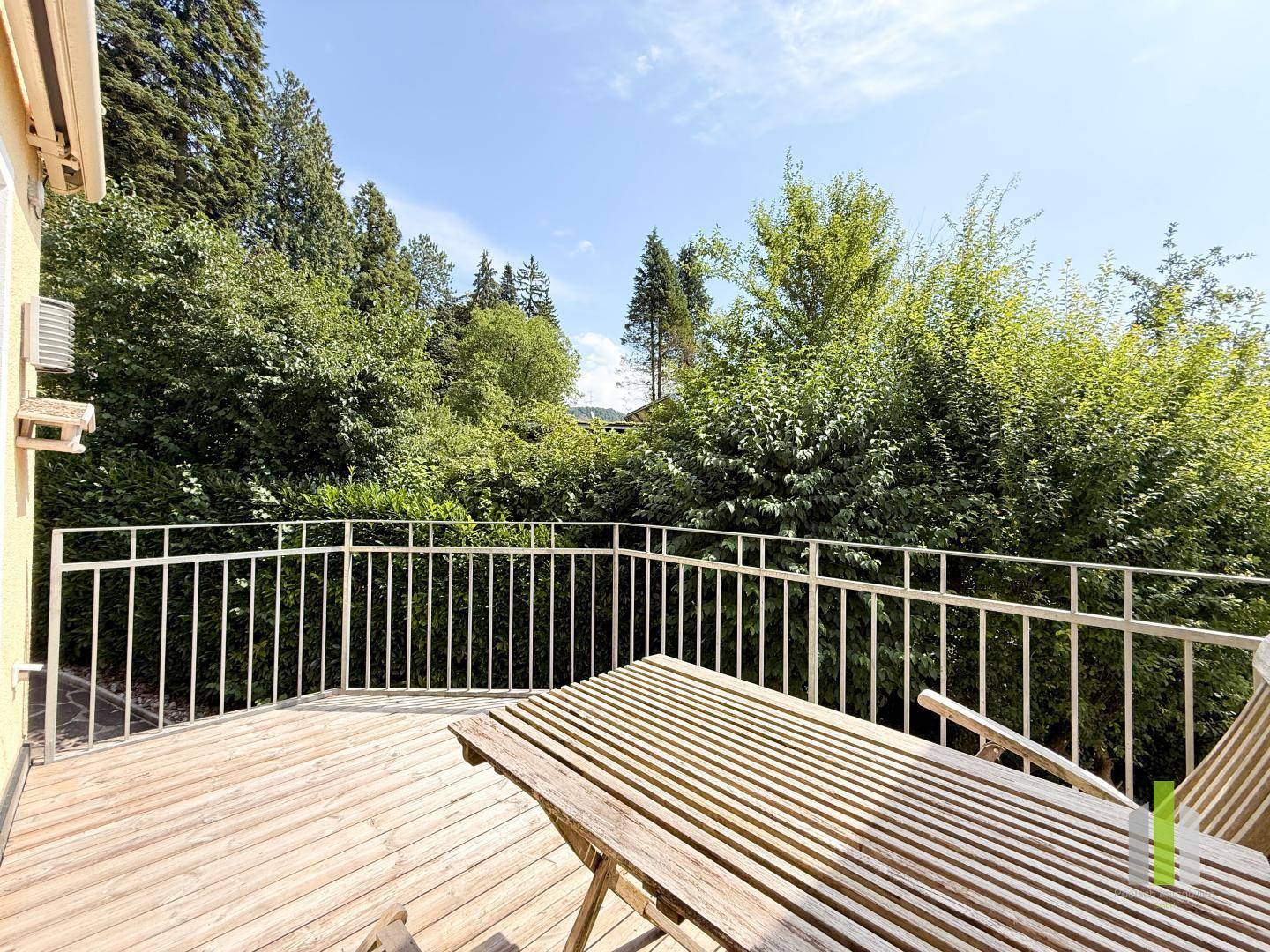 Balkon mit Holzboden, Gartentisch und umgeben von üppigem Grün unter blauem Himmel.