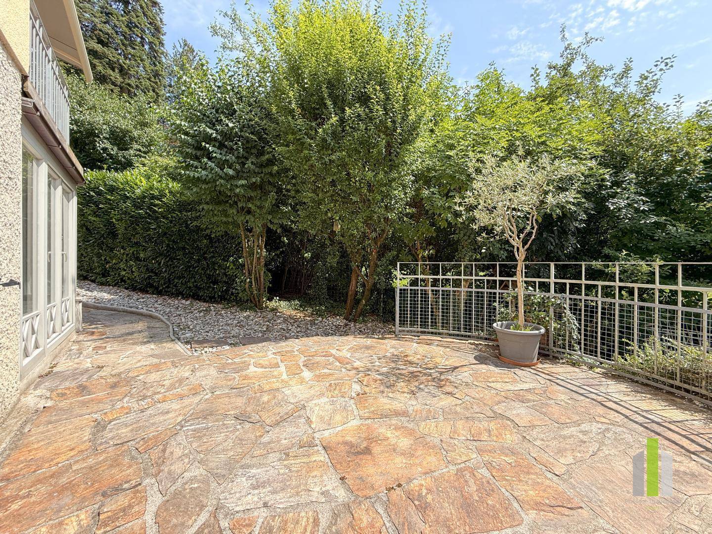 Große gepflasterte Terrasse mit Zugang zum Garten und umgeben von hohen Hecken.