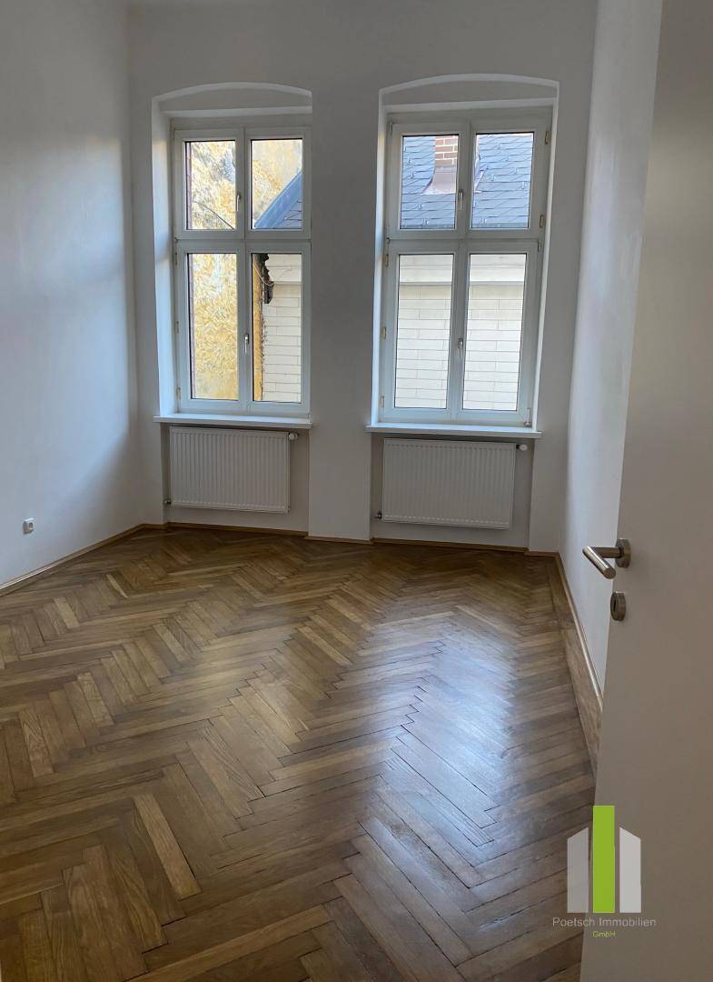 Gemütliches Zimmer mit Fischgrätparkett, zwei Fenstern und Heizkörpern unter den Fenstern.