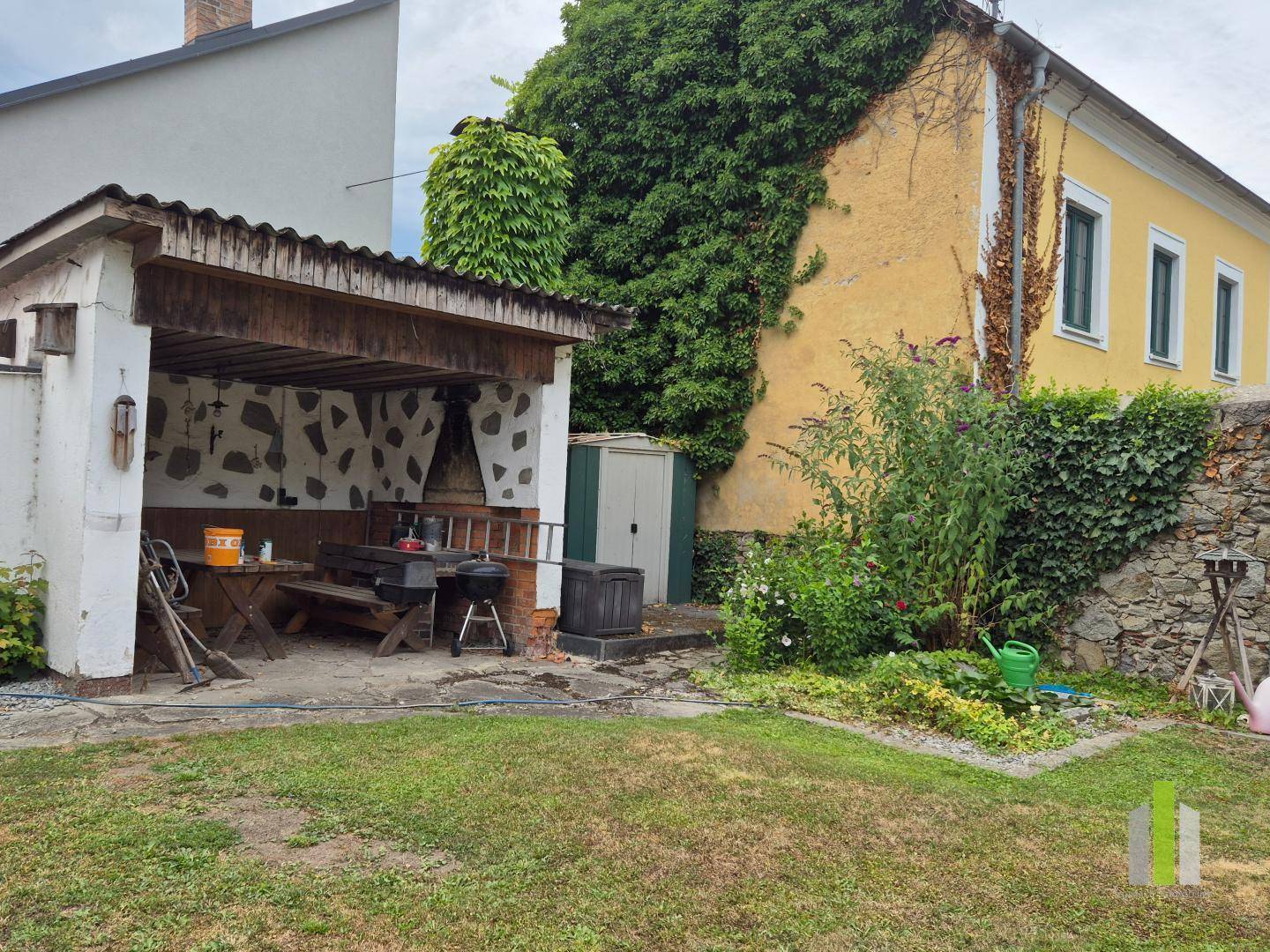Grüner Gartenbereich mit überdachtem Grillplatz, Holztisch und einem kleinen Geräteschuppen.