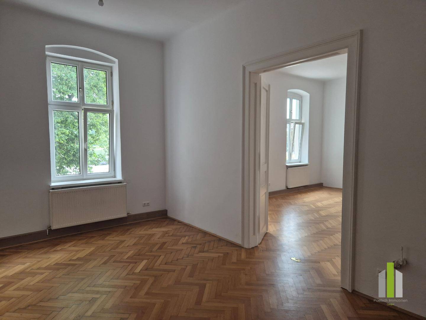 Geräumiges Zimmer mit Fischgrätparkett und Blick durch eine offene Tür in einen weiteren Raum.