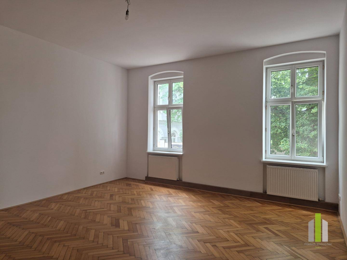 Geräumiges Zimmer mit Fischgrätparkett, hohen Fenstern und weiß gestrichenen Wänden.