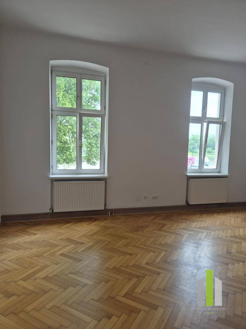 Helles Zimmer mit Fischgrätparkettboden, zwei hohen Fenstern und Blick ins Grüne.