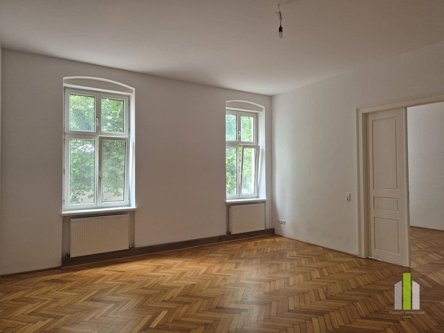 Helles Zimmer mit Fischgrätparkett und einer breiten Tür, die zu einem weiteren Raum führt.